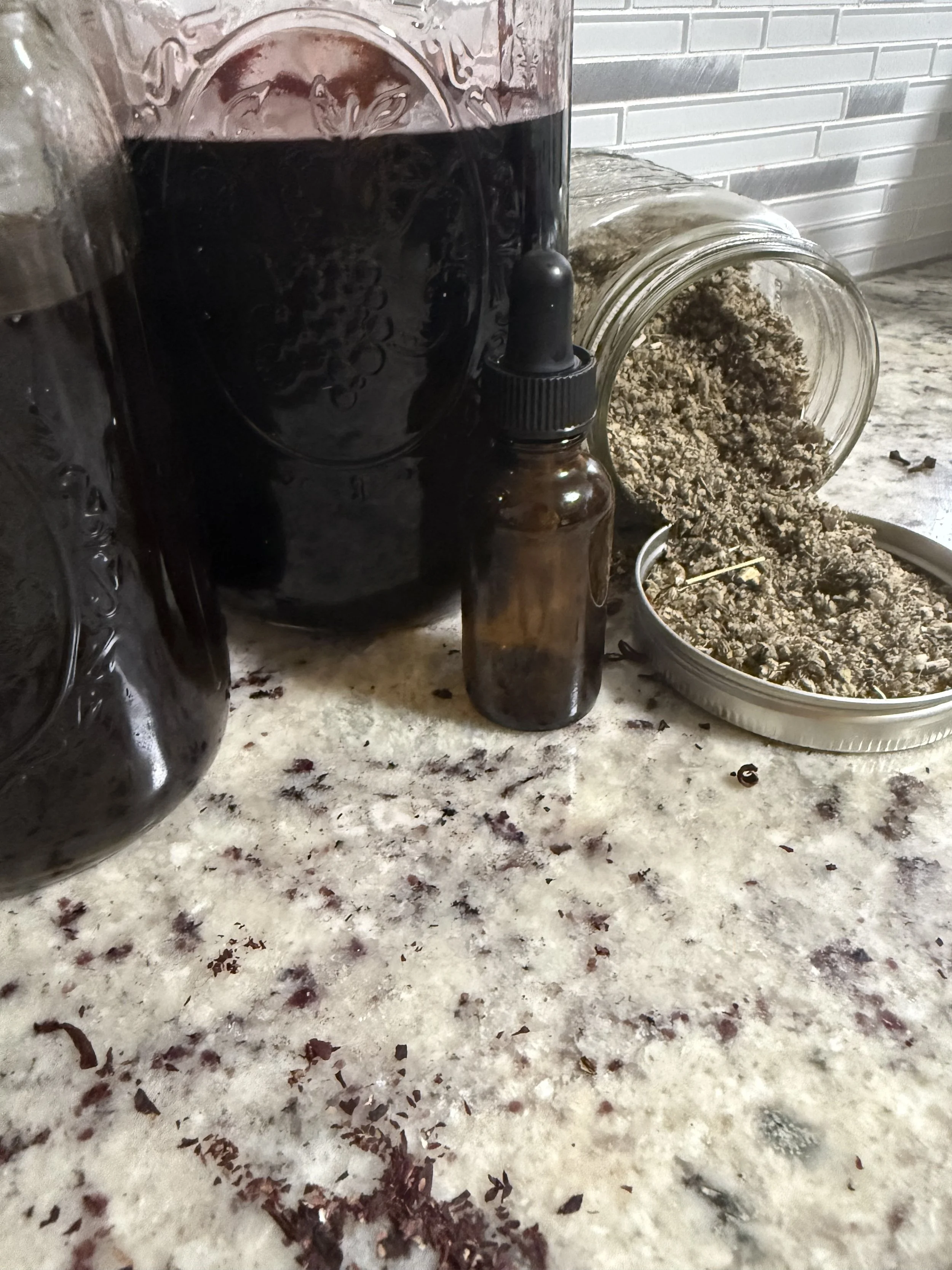 Open glass jar with dried herbs or plant material spilled on a speckled countertop, with a small dark amber dropper bottle nearby.