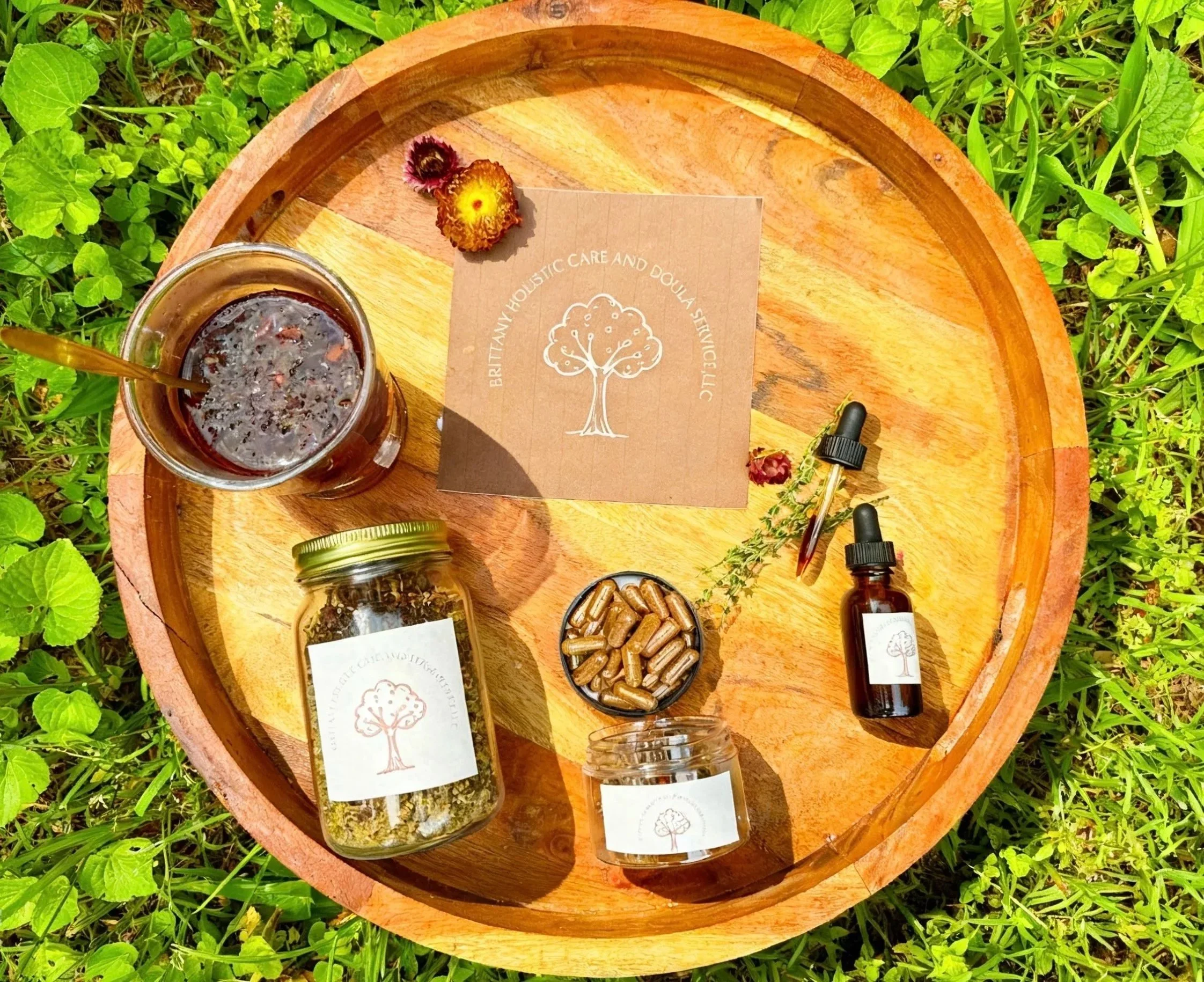 A round wooden tray on green grass holds various herbal products, including a jar of dried herbs, a small bowl of capsules, a small dropper bottle, a small jar, and a glass with a dark beverage. A brown paper with a tree logo and