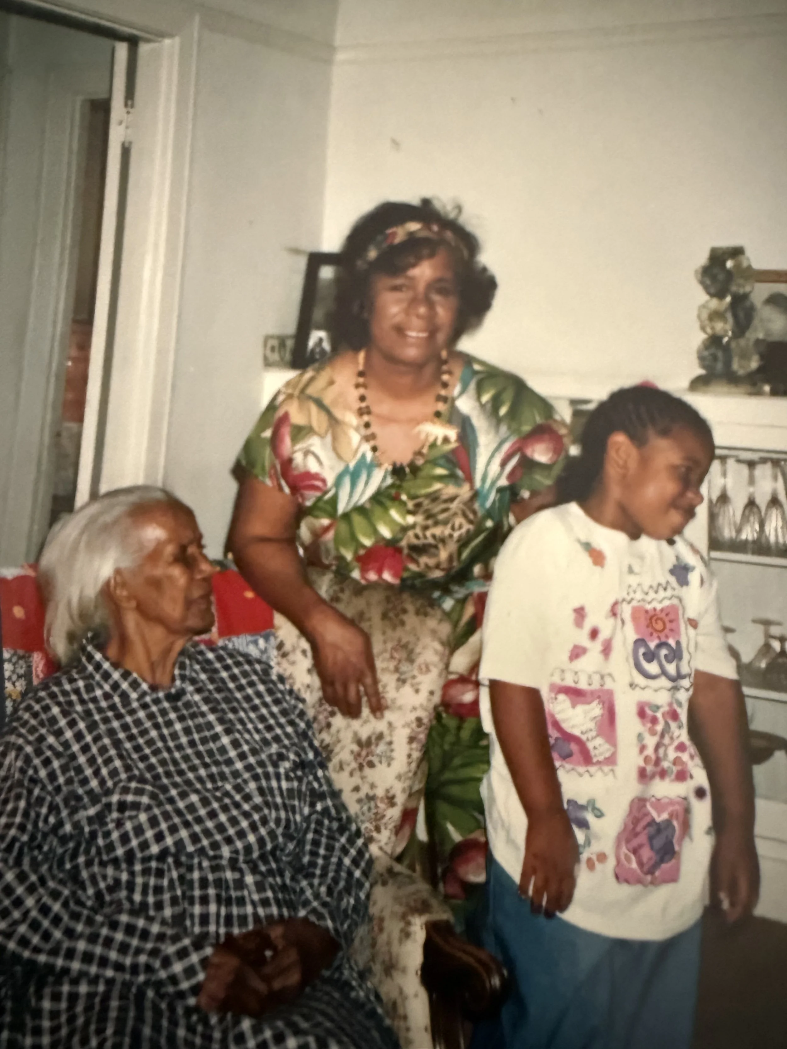 Three women in a living room, one sitting with gray hair wearing a checkered dress, and two standing, one with short curly hair wearing a floral shirt and the other with braided hair wearing a colorful shirt with patches.