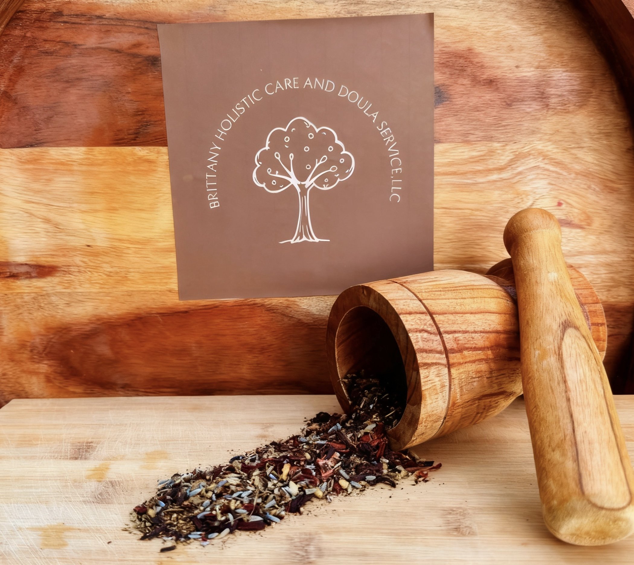 A wooden scoop tipped over with loose herbal tea blend spilling onto a wooden surface, with a card reading 'Britanny Holistic Care and Doula Service,' and wooden rolling pin nearby.