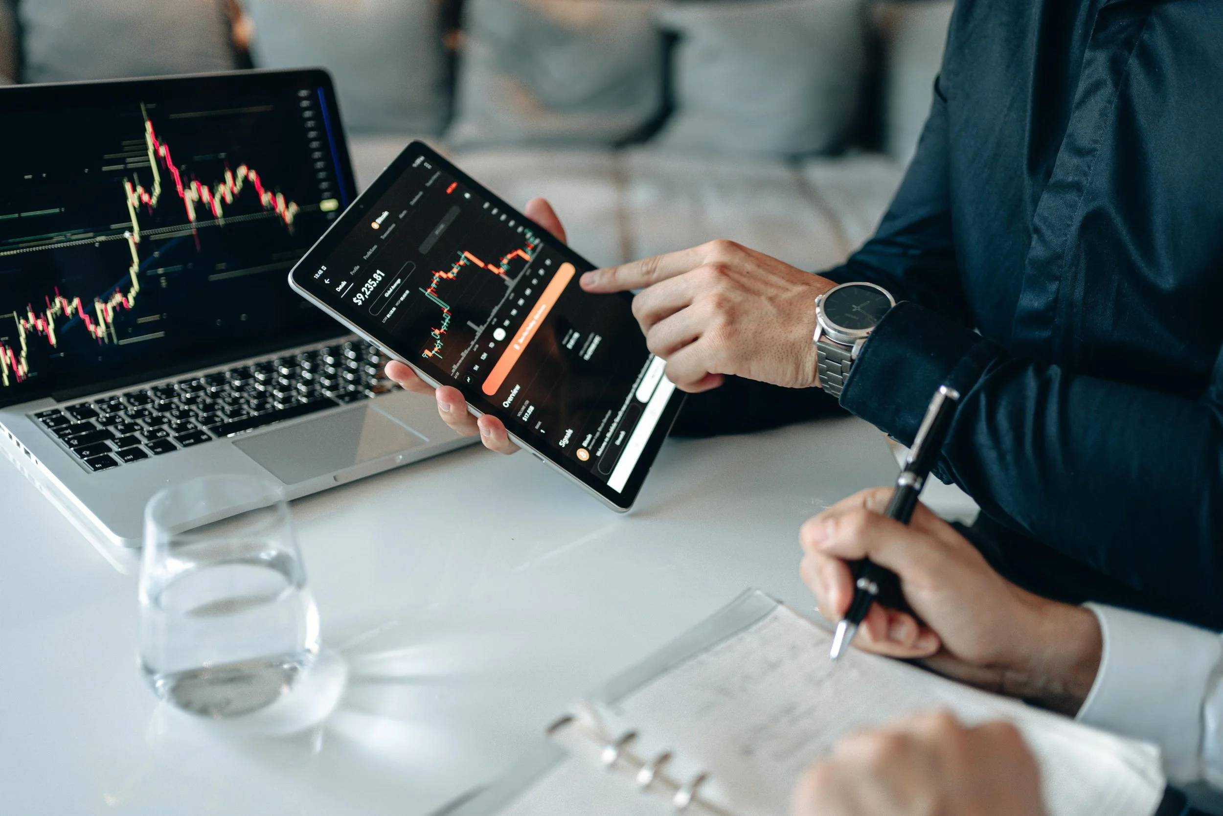 Person analyzing stock market charts on a digital tablet and a laptop with financial graphs, with a glass of water and notes on the white desk.