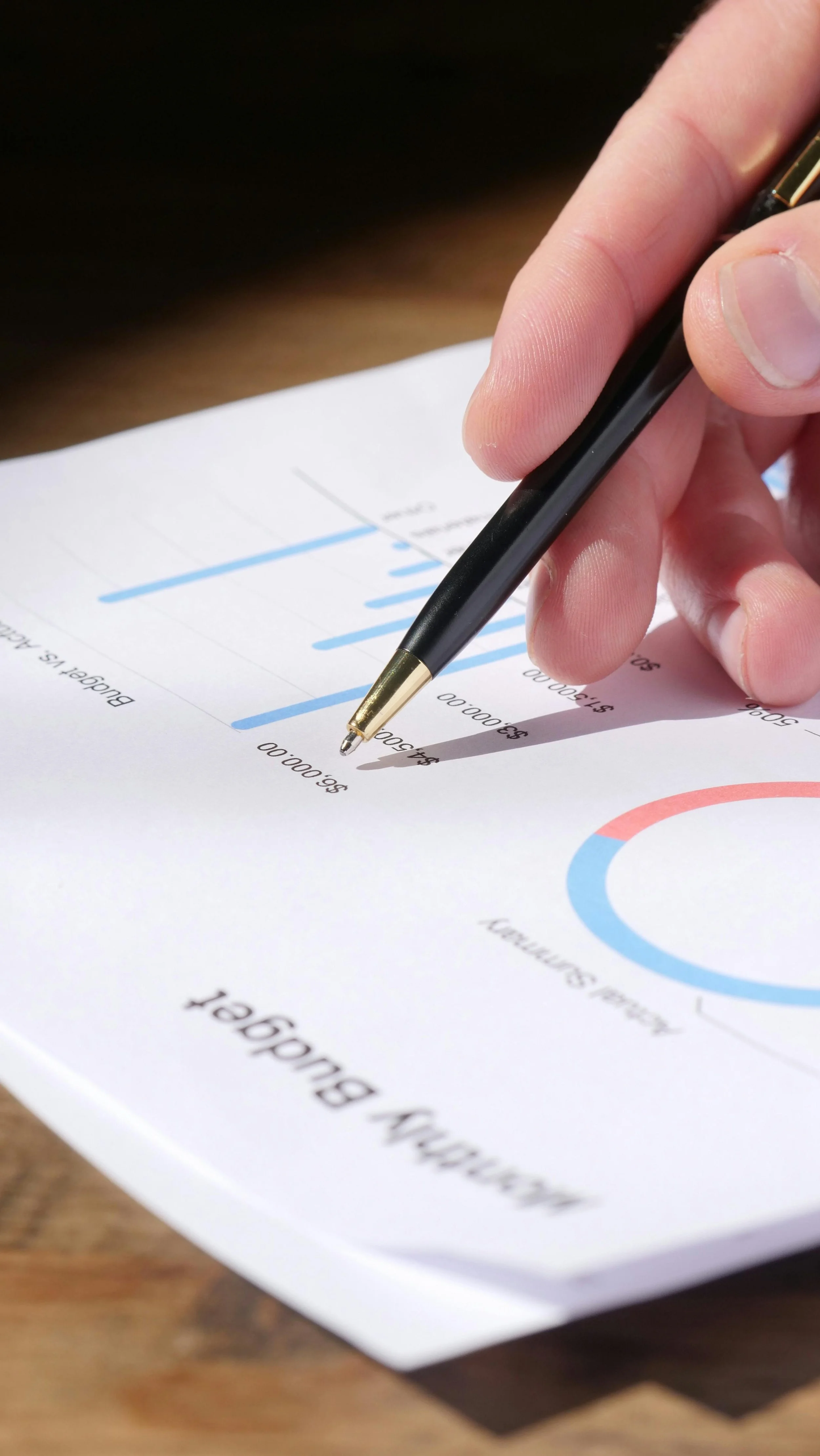 A person holding a black and gold pen is pointing at a line graph on a business report or presentation sheet with colorful charts and text.