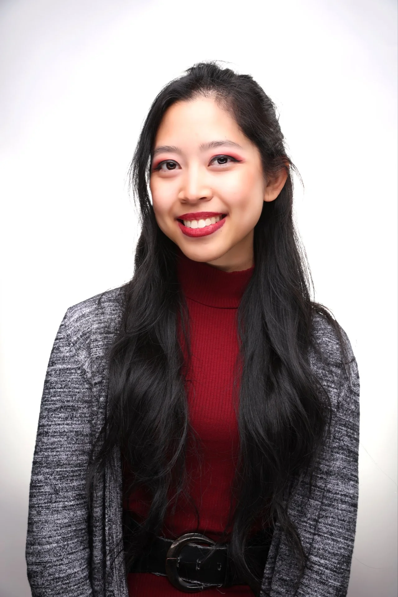 Portrait of a young Asian woman with long black hair, wearing a red mockneck dress and a gray cardigan, smiling against a white background.