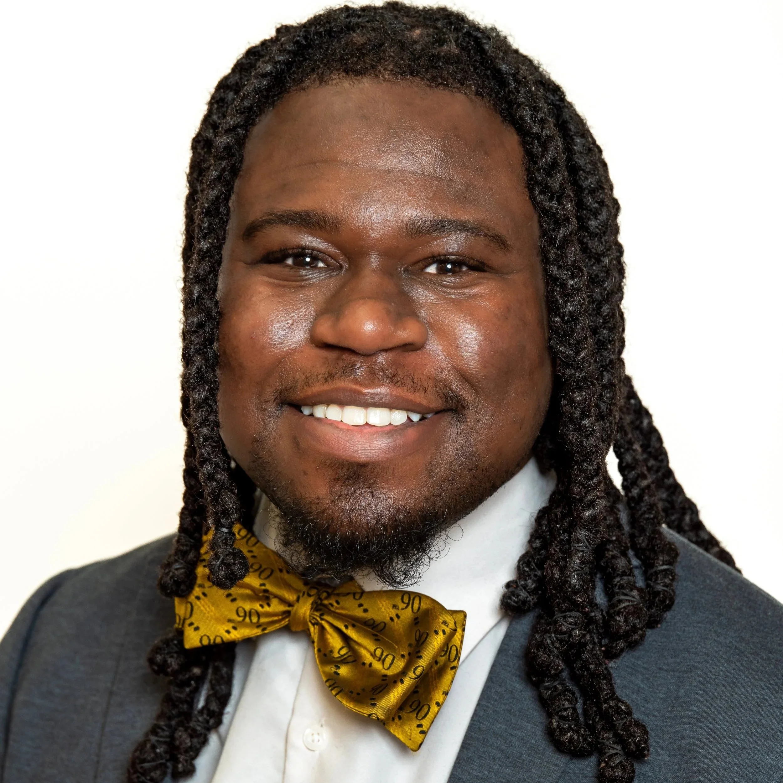 Portrait of a smiling man with dreadlocks, wearing a suit and a yellow bow tie with black numbers, on a white background.