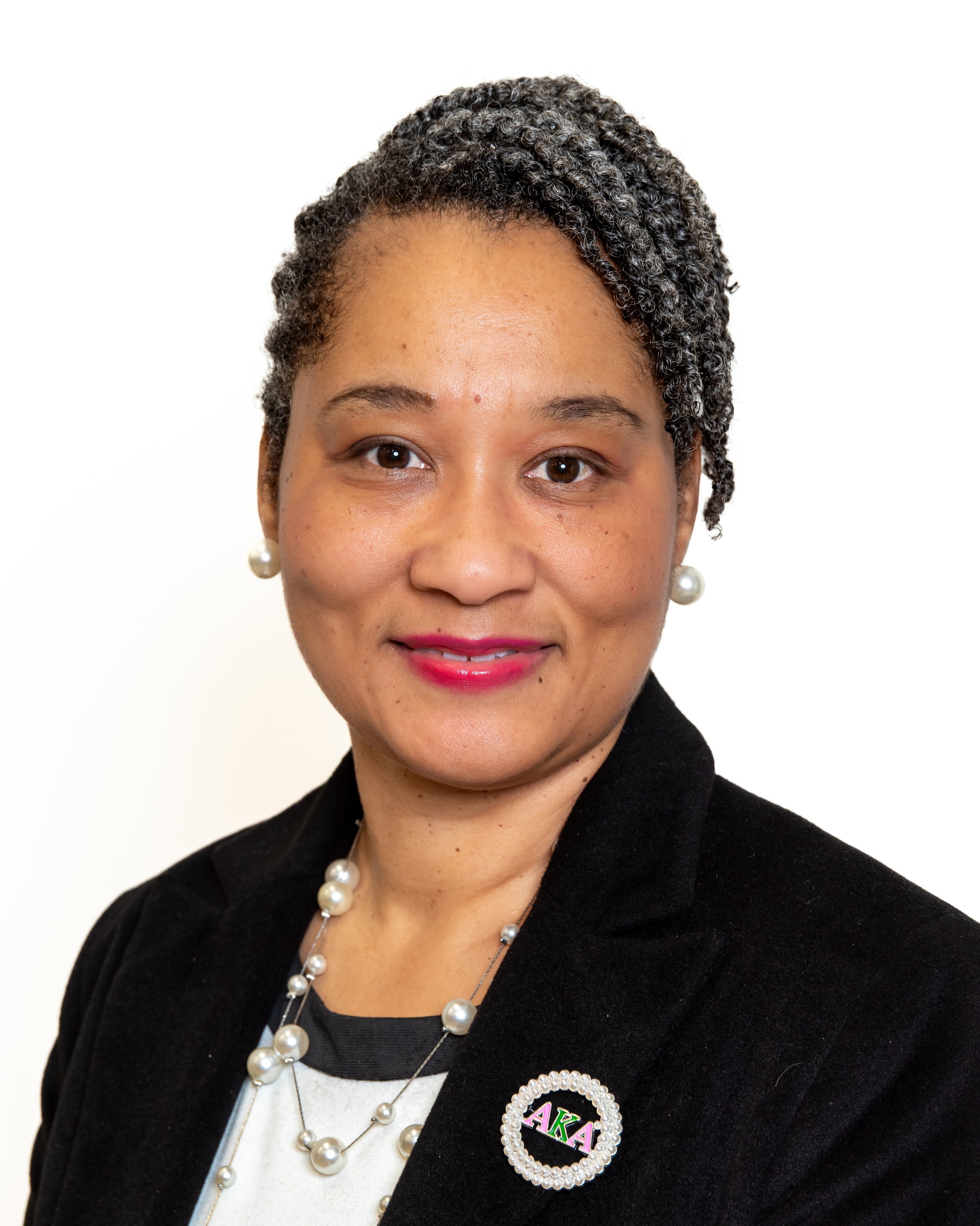 Portrait of a woman with short, curly gray hair, wearing pearl earrings and necklace, a black blazer, a polka-dot shirt, and an AKA pin, standing against a white background.