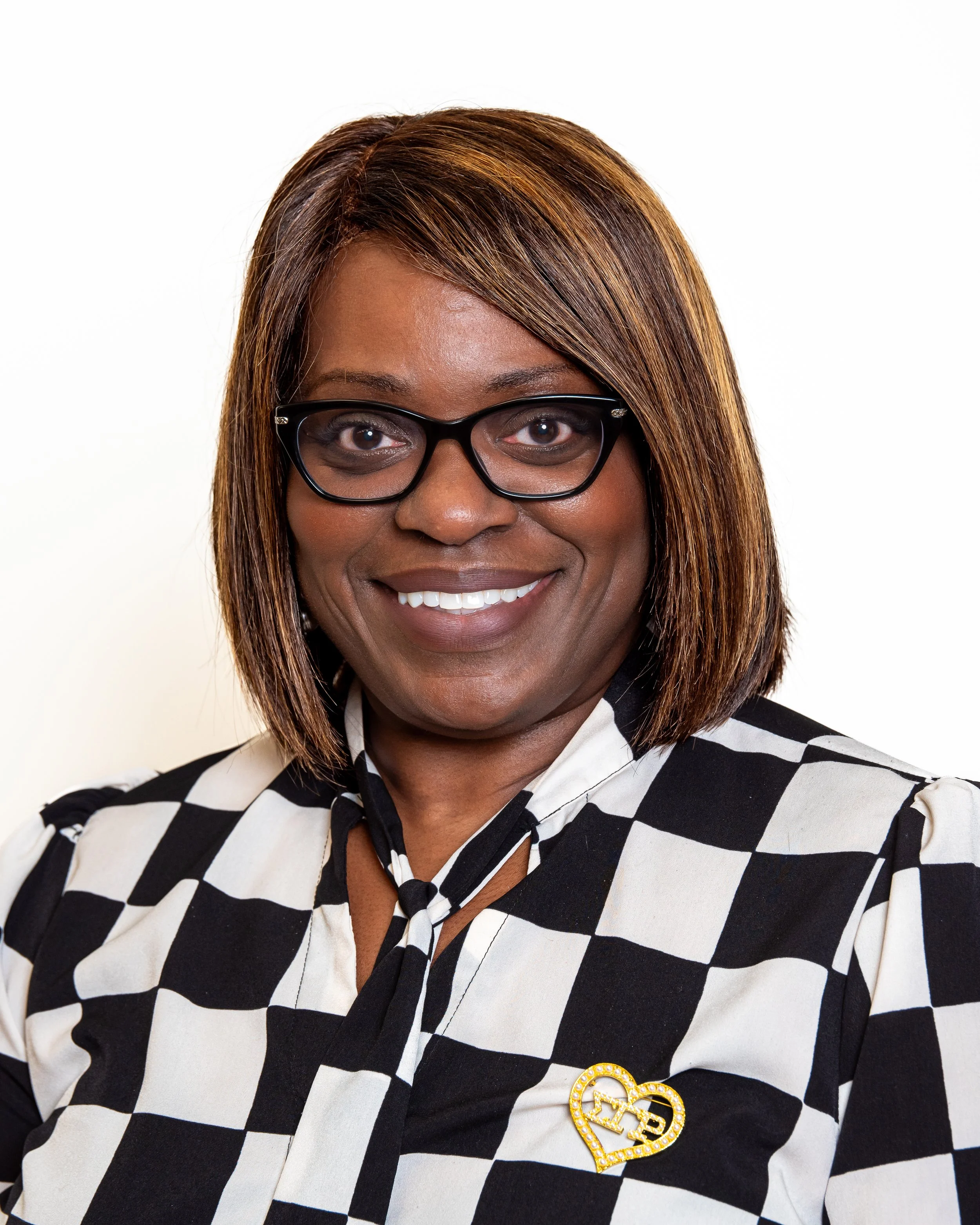A woman with medium-length brown hair with highlights and glasses, smiling, wearing a black and white checkered blouse with a decorative bow tie and a gold heart pin.