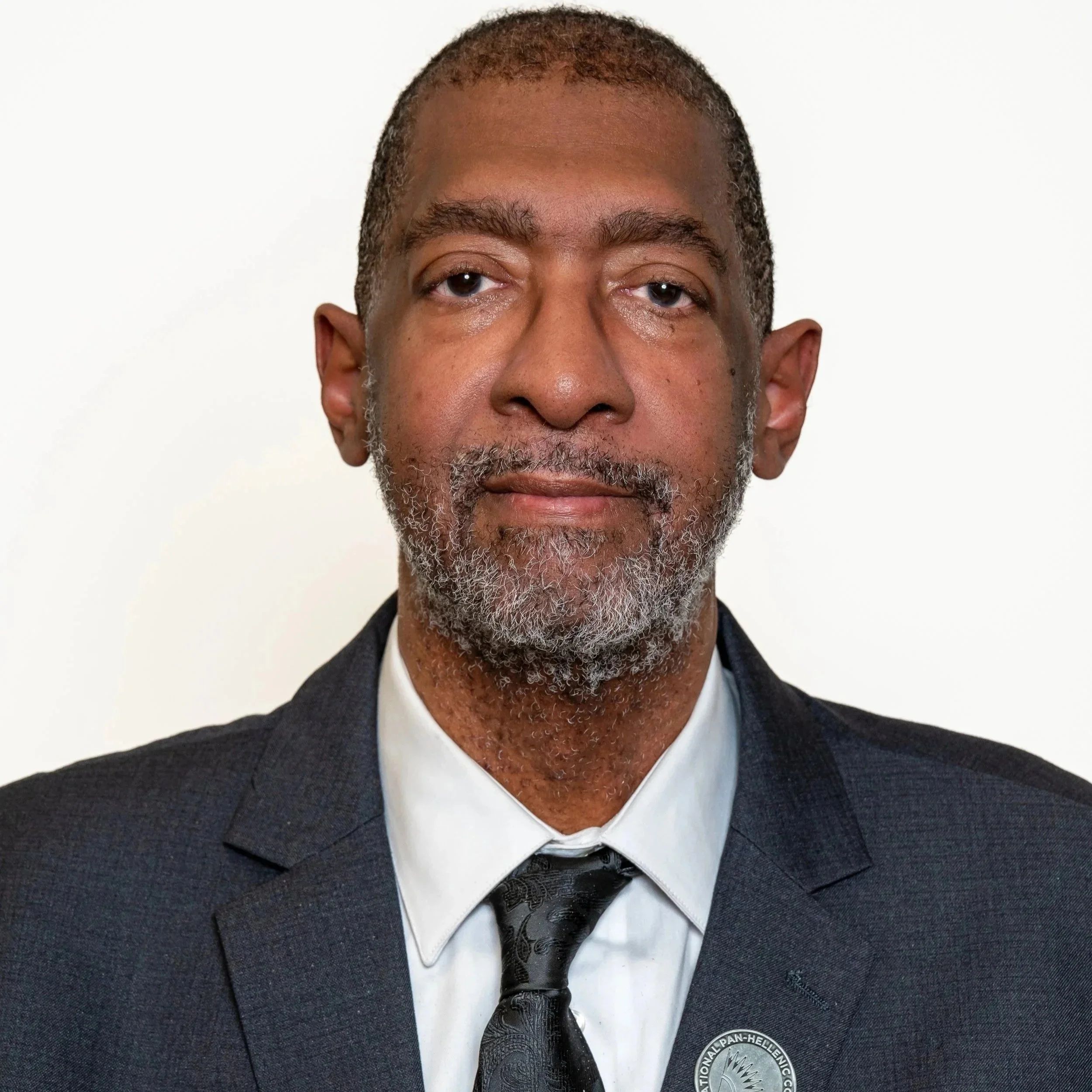 A close-up portrait of an older African American man with gray hair and beard, wearing a dark suit, white shirt, and patterned black tie, against a plain white background.