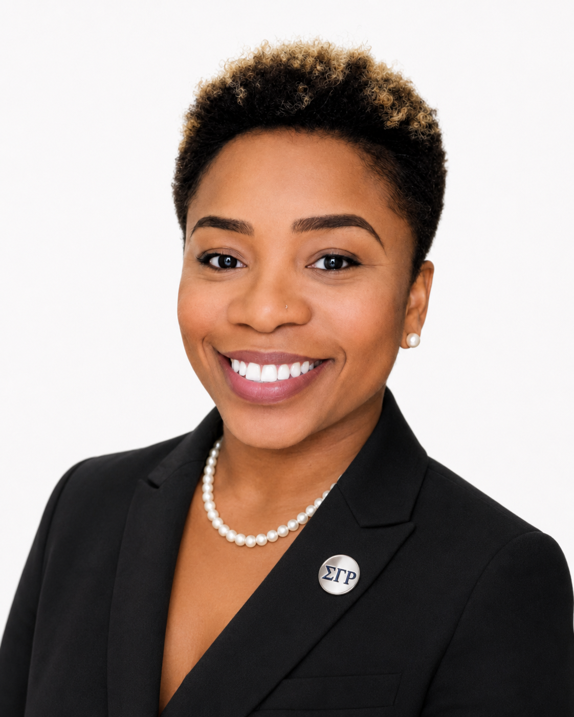 A woman with dark blonde dreadlocks, wearing a black headband, red lipstick, a black checkered blazer with a Sigma Theta Pi brooch, and a white shirt, smiling against a white background.