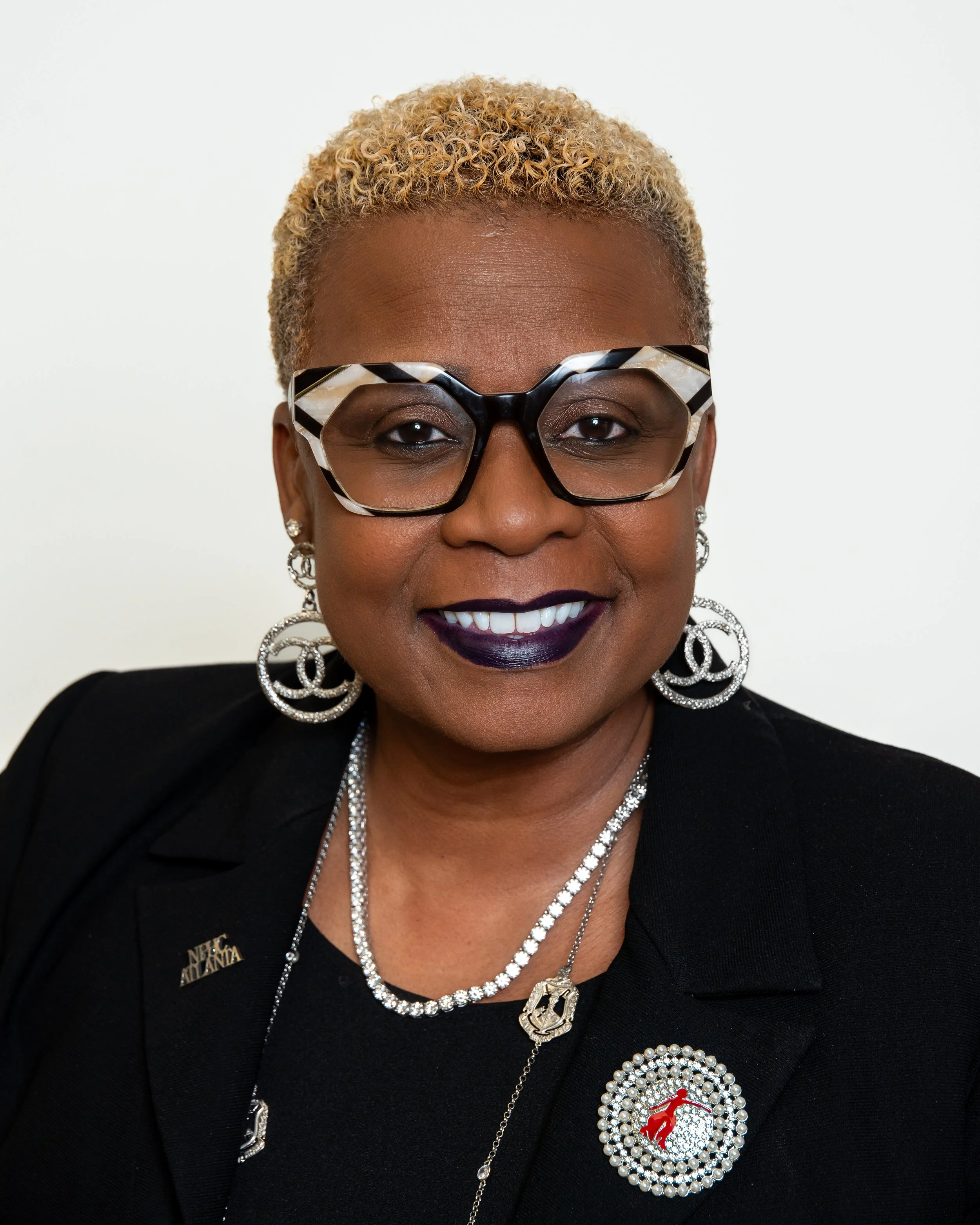 Close-up portrait of an African American woman with short, curly, blonde hair, wearing black and white patterned glasses, large silver earrings, a pearl necklace, and a black blazer with a decorative pin. She has dark lipstick and is smiling at the camera against a plain white background.