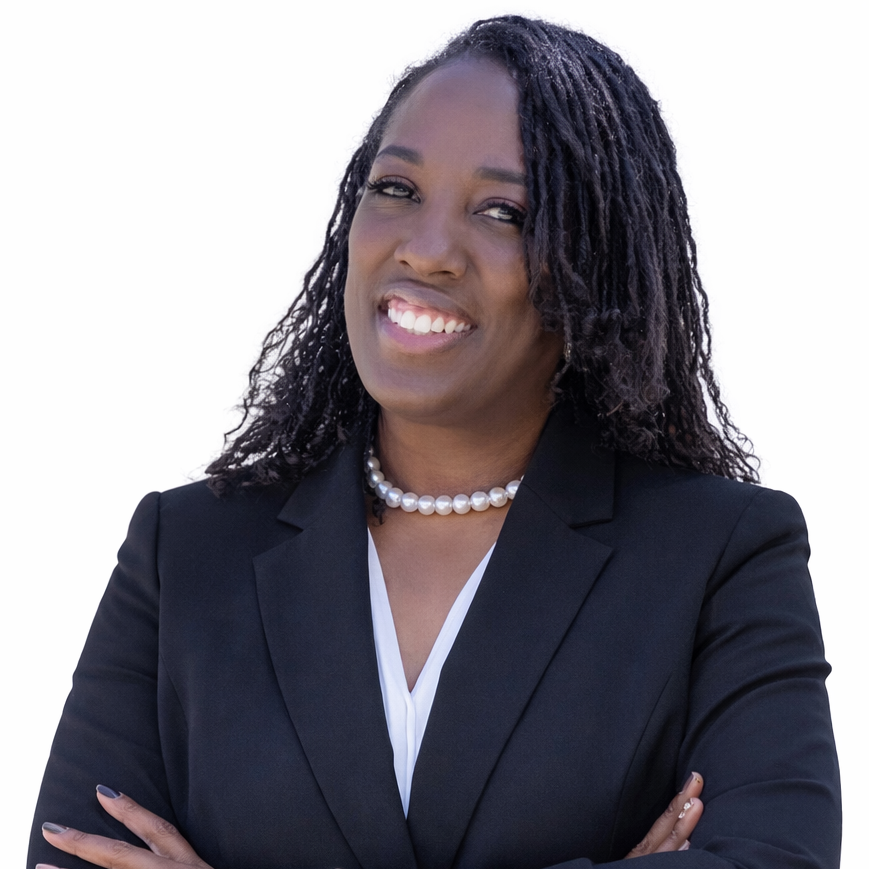 A woman in a black blazer with arms crossed, smiling, wearing a pearl necklace, against a white background.
