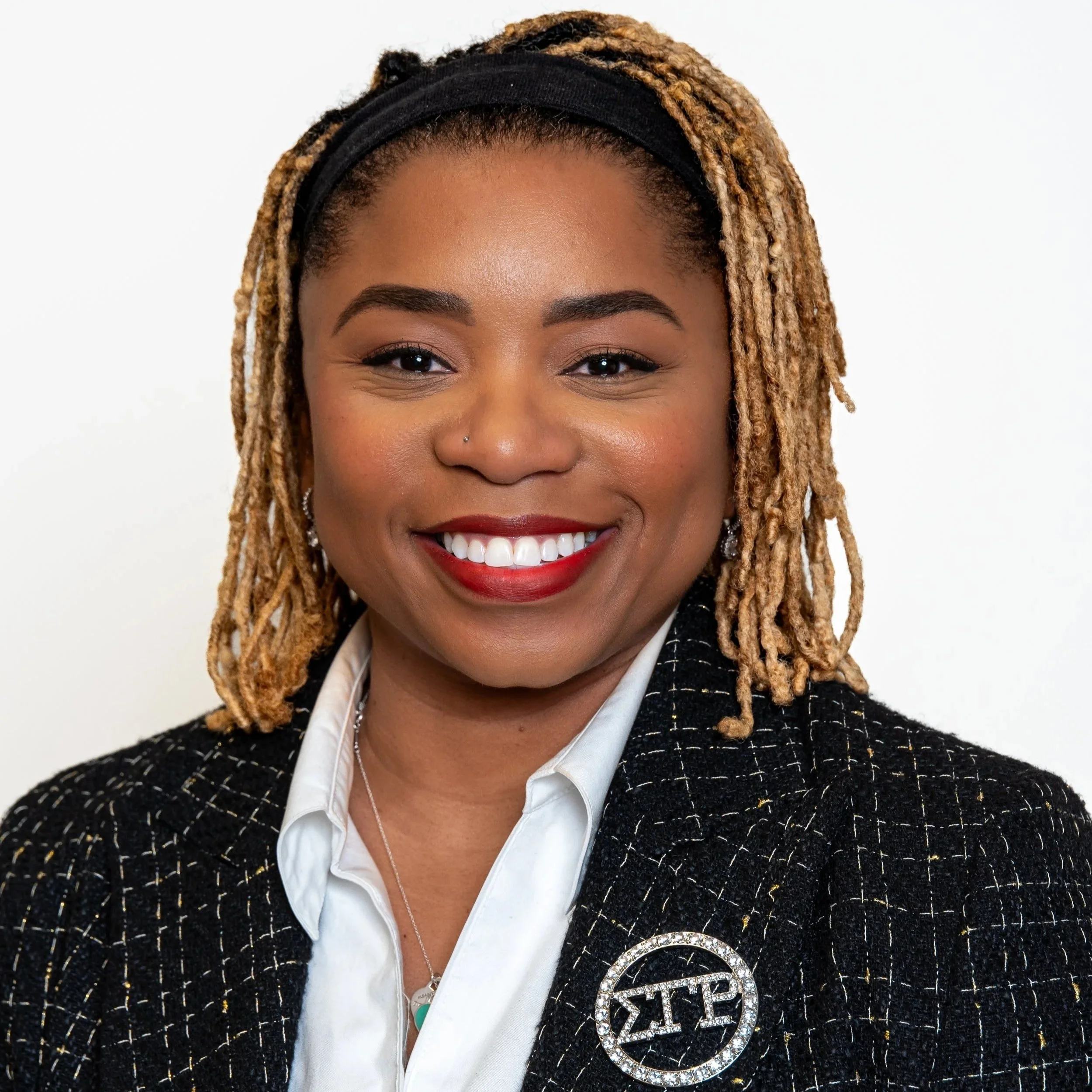 A woman with dark blonde dreadlocks, wearing a black headband, red lipstick, a black checkered blazer with a Sigma Theta Pi brooch, and a white shirt, smiling against a white background.
