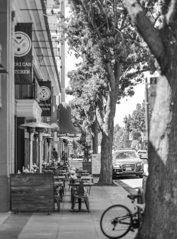 Street scene with outdoor seating, trees, storefronts, and a parked car on a sunny day.