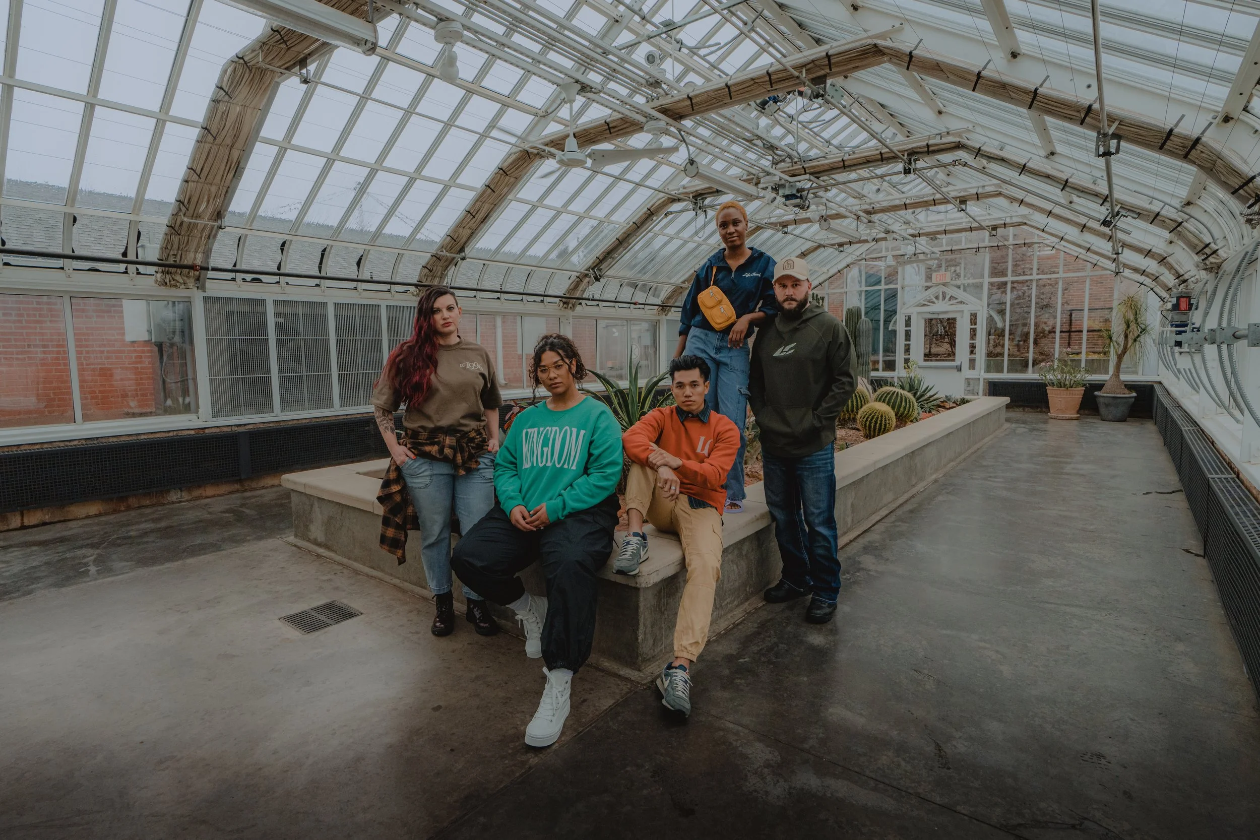 Five diverse young adults inside a greenhouse, with a concrete bench and cactus plants, posing for a group photo.