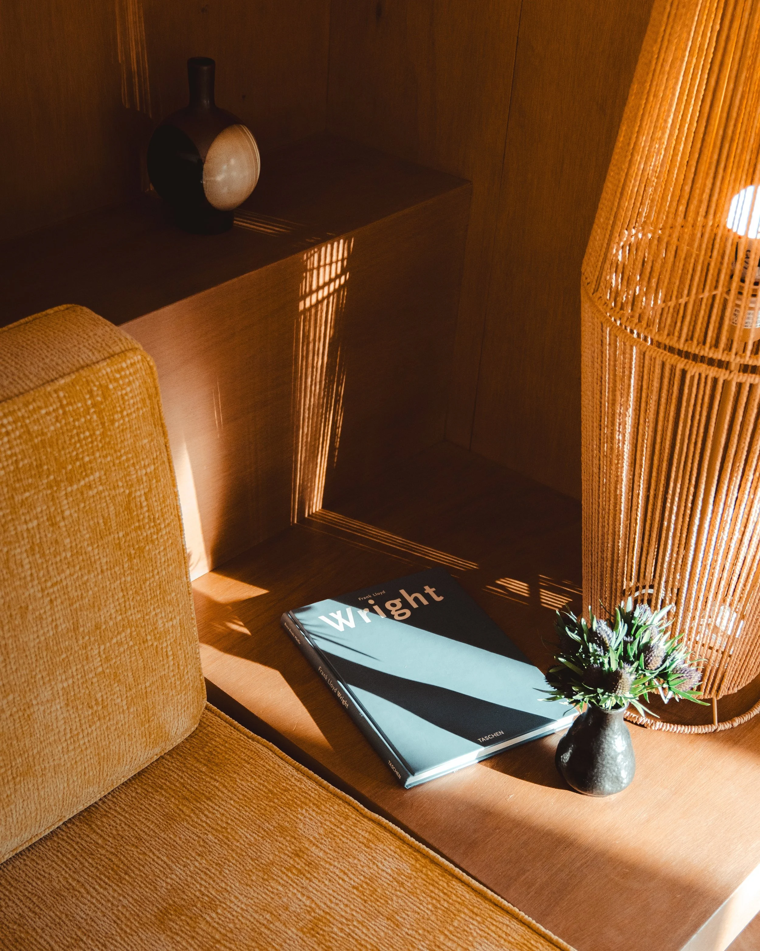 A bedside table with a book titled 'Wright,' a small black vase with flowers, and a wooden lamp, illuminated by sunlight casting shadows.