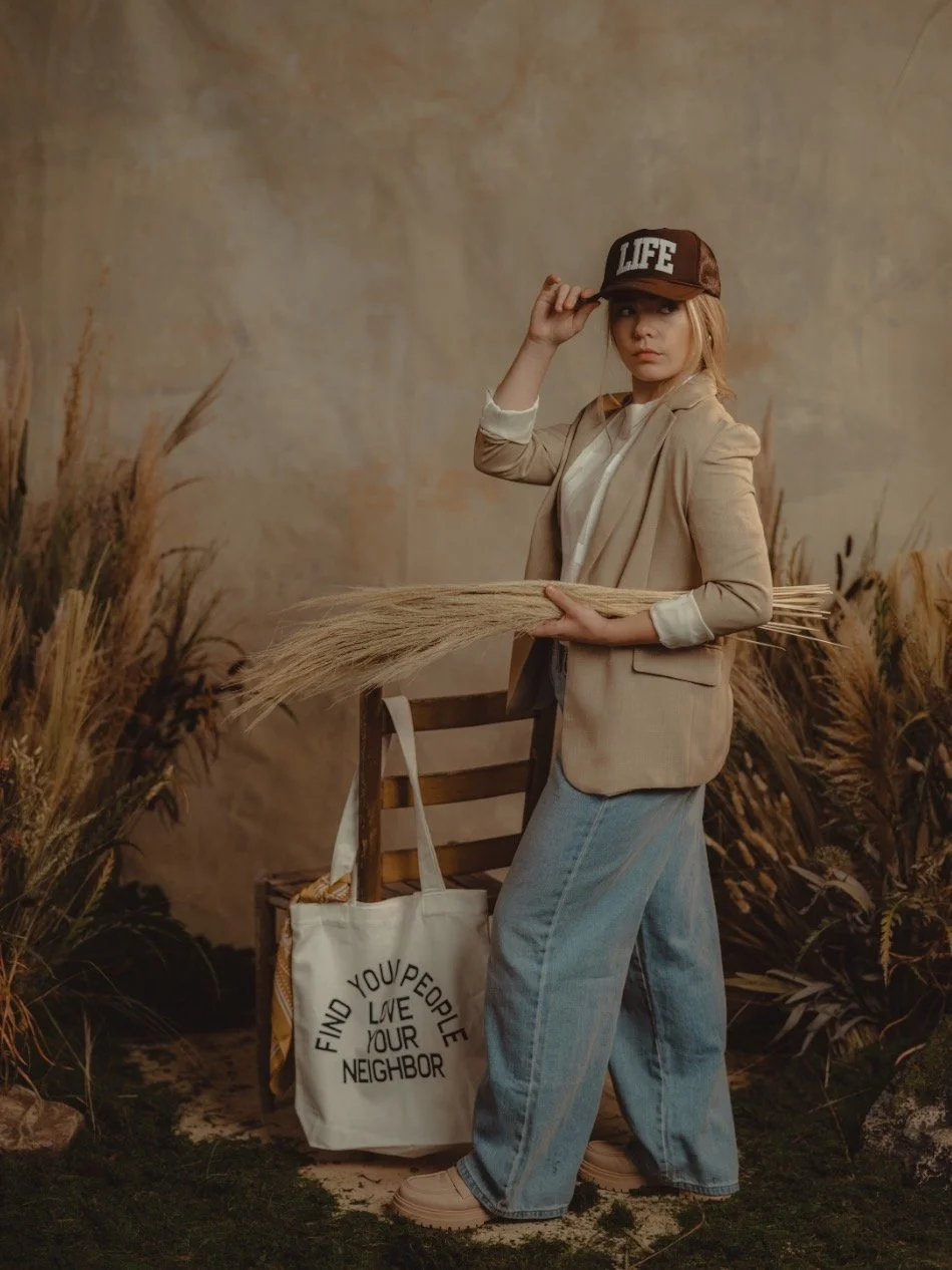 A woman wearing a beige blazer, light blue jeans, a white shirt, and a brown cap that says 'LIFE'. She is holding a bundle of wheat and standing next to a chair with a white tote bag that says 'FIND YOUR NEIGHBOR, LOVE YOUR NEIGHBOR'. The background 