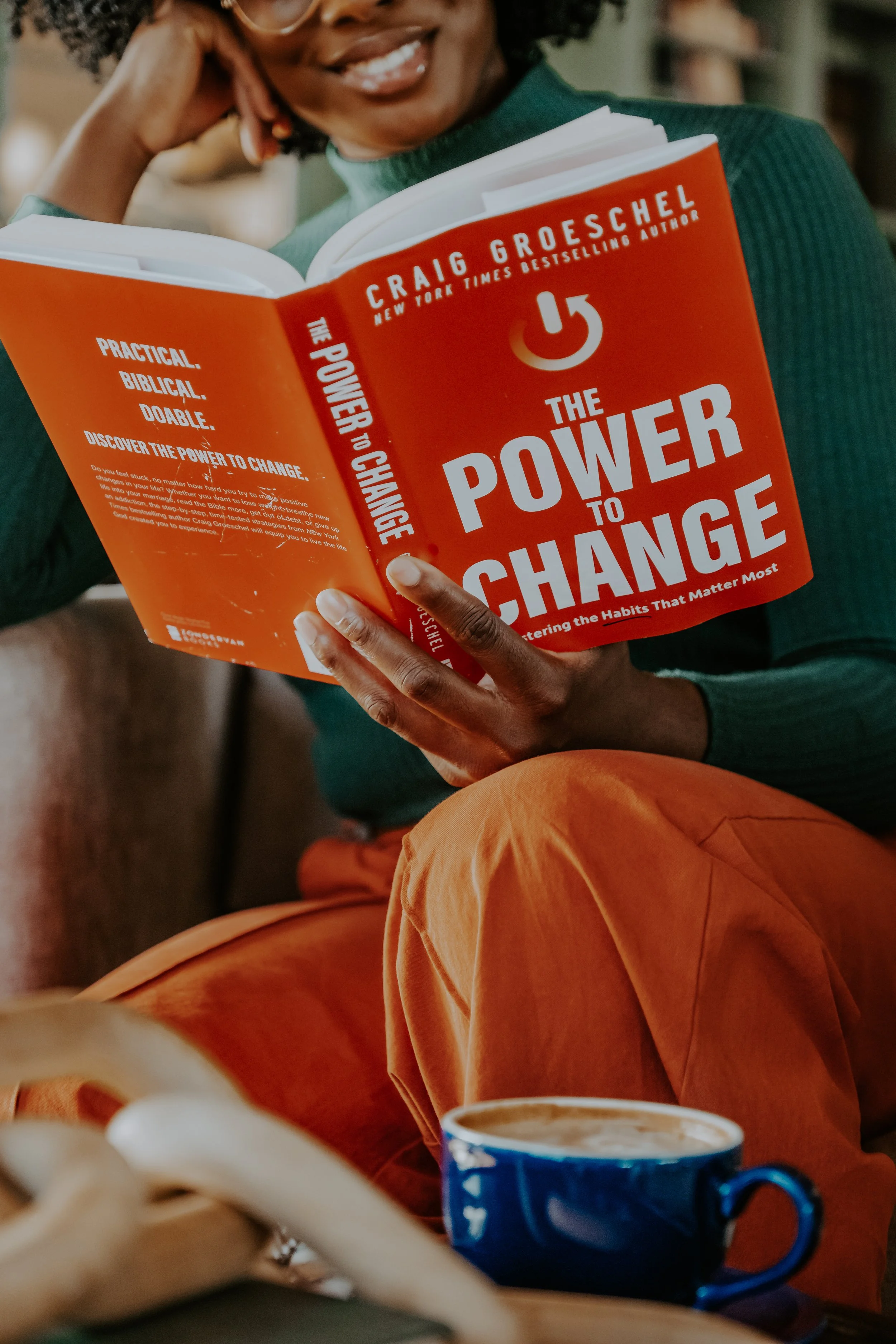 A woman sitting on a couch, holding and reading a book titled 'The Power to Change' by Craig Groeschel. She is smiling, wearing an orange pair of pants and a green sweater. There is a blue cup of coffee or tea in front of her on the table.