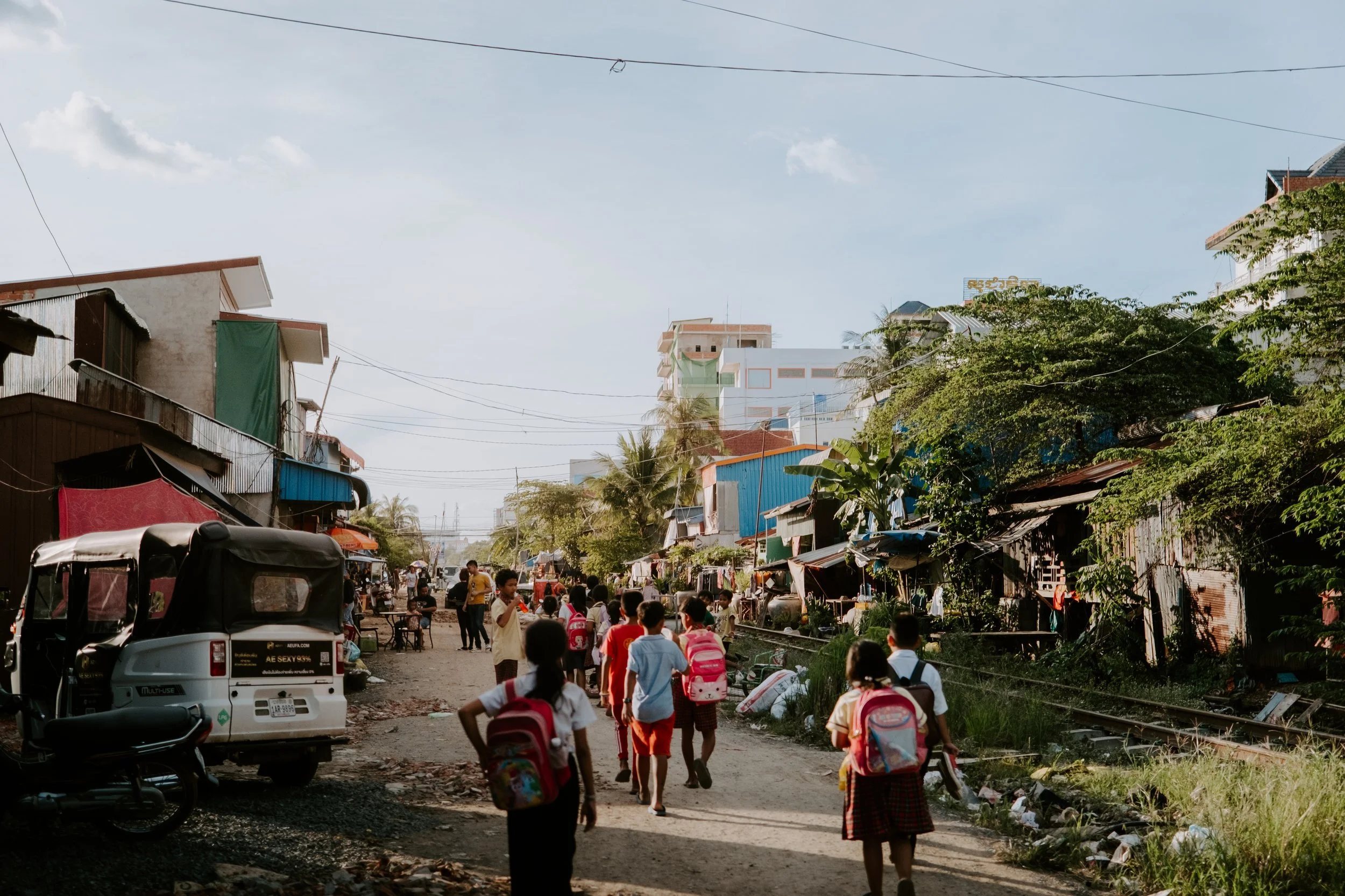 Children walking on a dirt street in a neighborhood with makeshift houses, some shops, and trees against a blue sky with a few clouds.