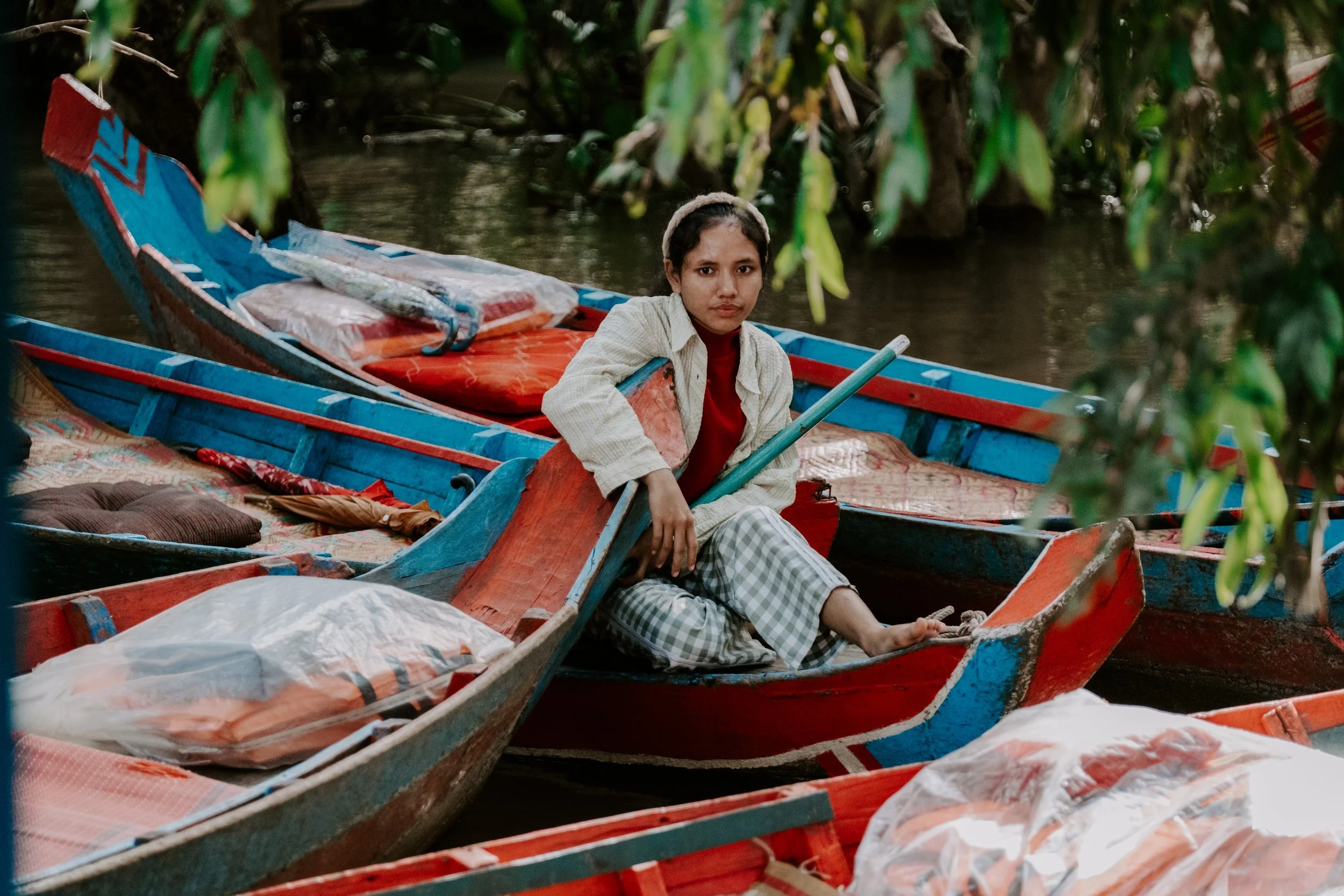 A young woman sitting in a small colorful boat on a river, amidst other boats, surrounded by green foliage.