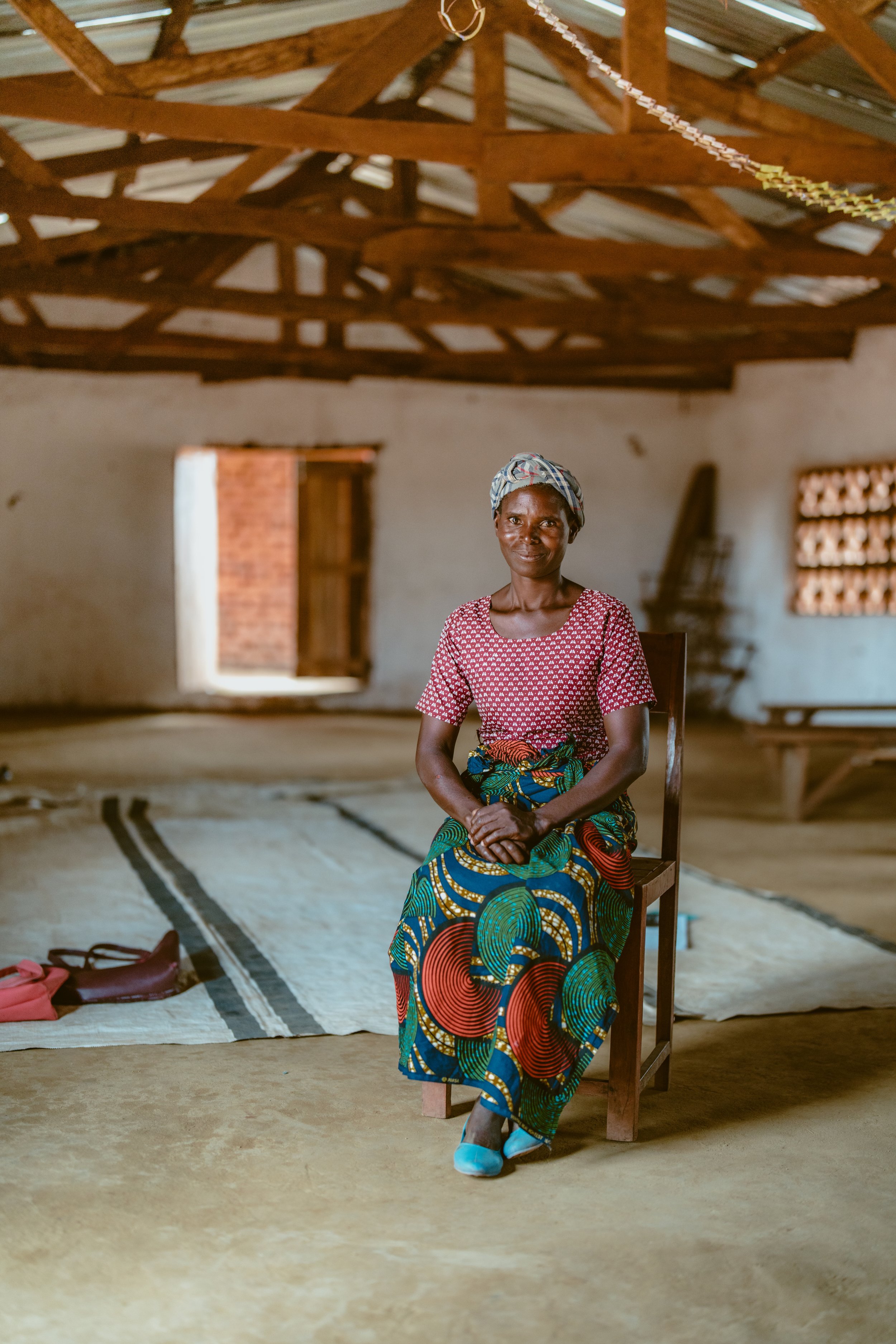 Woman sitting on a wooden chair inside a simple room with bare walls, a thatched roof, and a window, wearing colorful patterned clothing and a headwrap.
