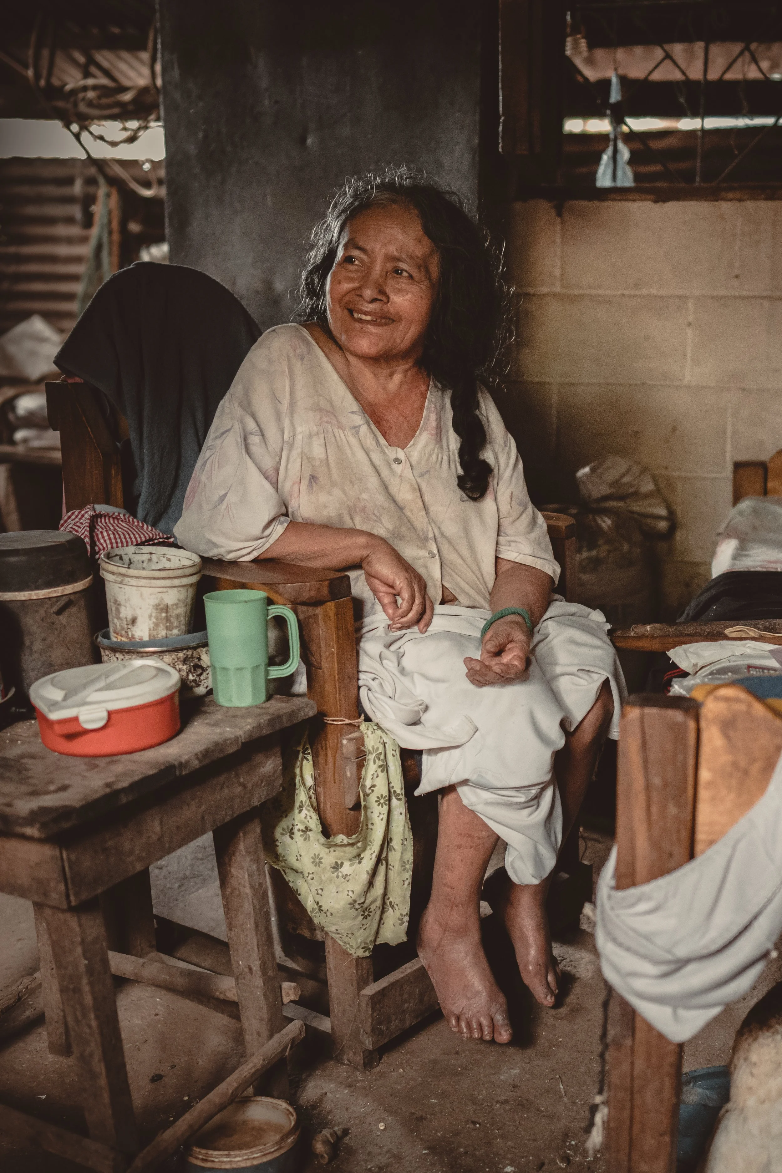 An older woman with dark, curly hair, wearing a light-colored blouse and skirt, sitting in a rustic, cluttered room, smiling and looking to her left.