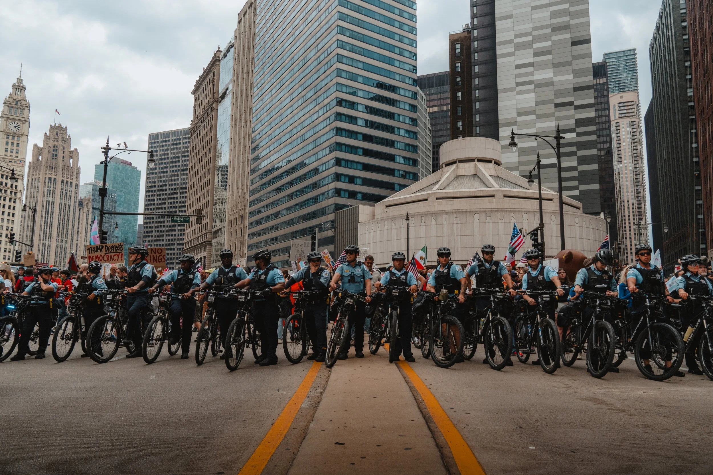 A line of police officers on bicycles blocking a city street during a protest, with protesters holding signs and flags in the background, skyscrapers and a church in the cityscape.