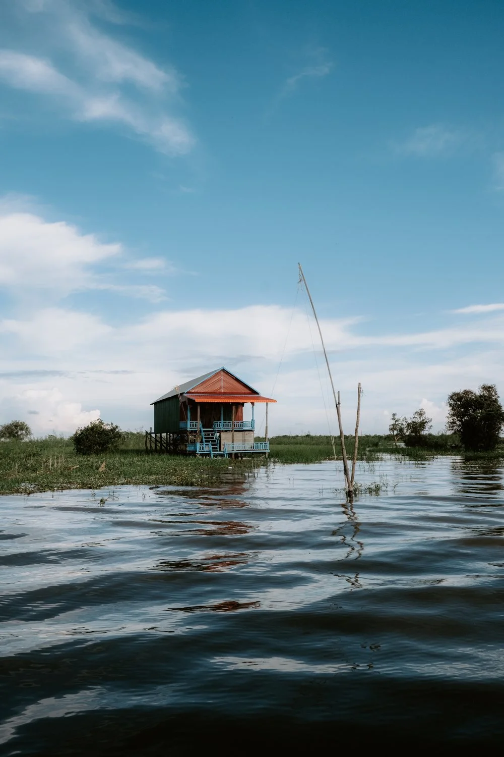A lakeside house with a thatched roof, blue railings, and a small staircase, located on a grass shore with calm water and a partly cloudy sky.