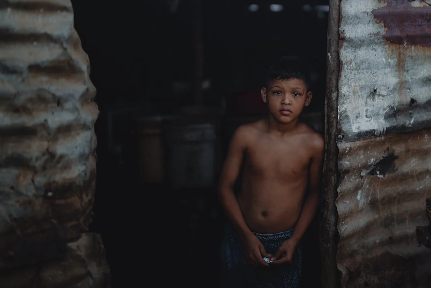 A young boy standing in a dark, rustic room, shirtless with a worried expression, holding something small in his hands, next to a corrugated metal wall.