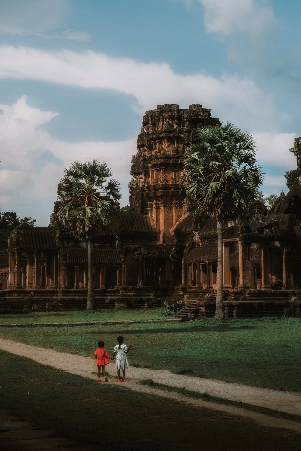 Two children walking on a dirt path in front of an ancient stone temple with tall palm trees and a partly cloudy sky.