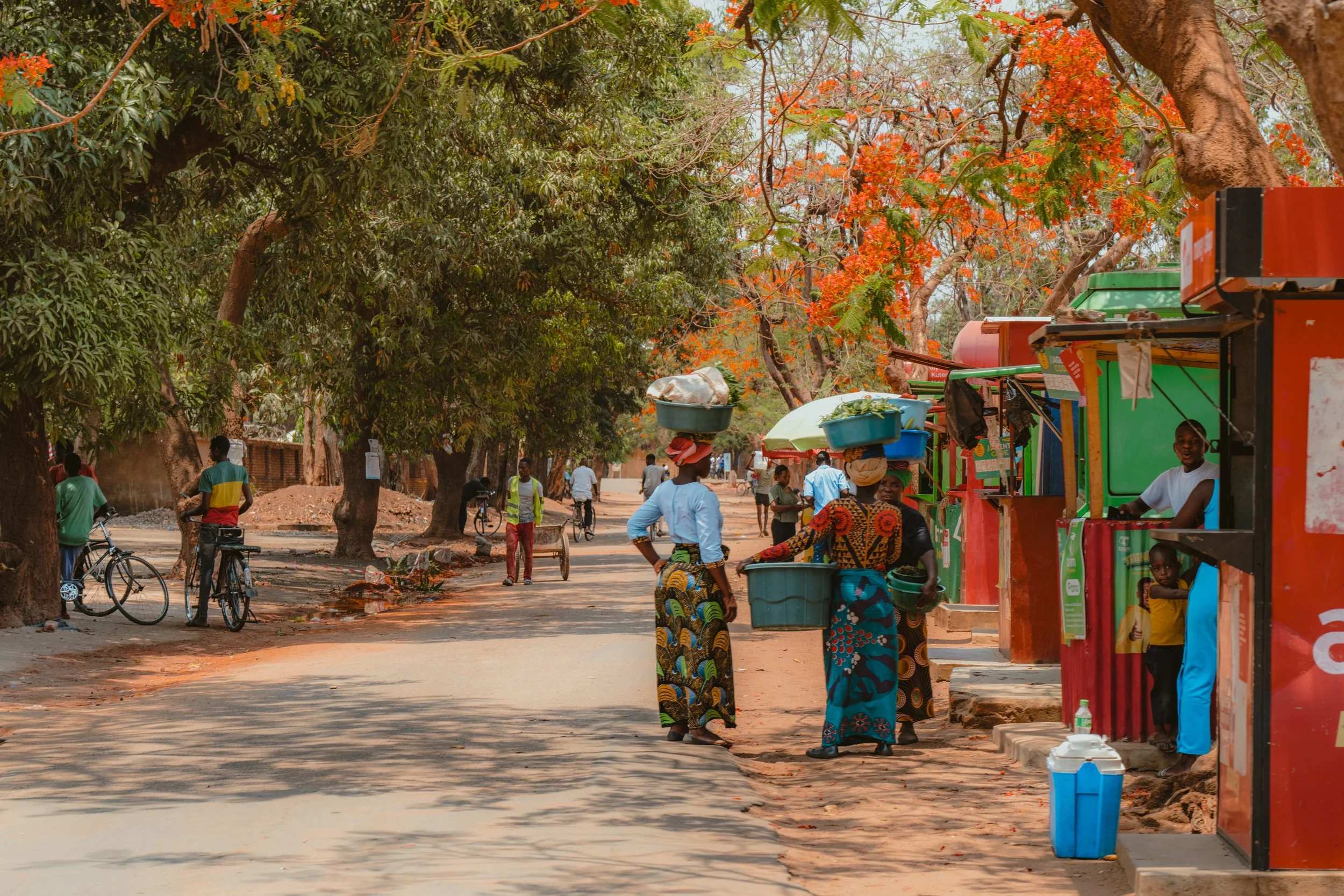 Street scene in a rural area with vendors selling goods, people riding bicycles, and trees providing shade on a sunny day.