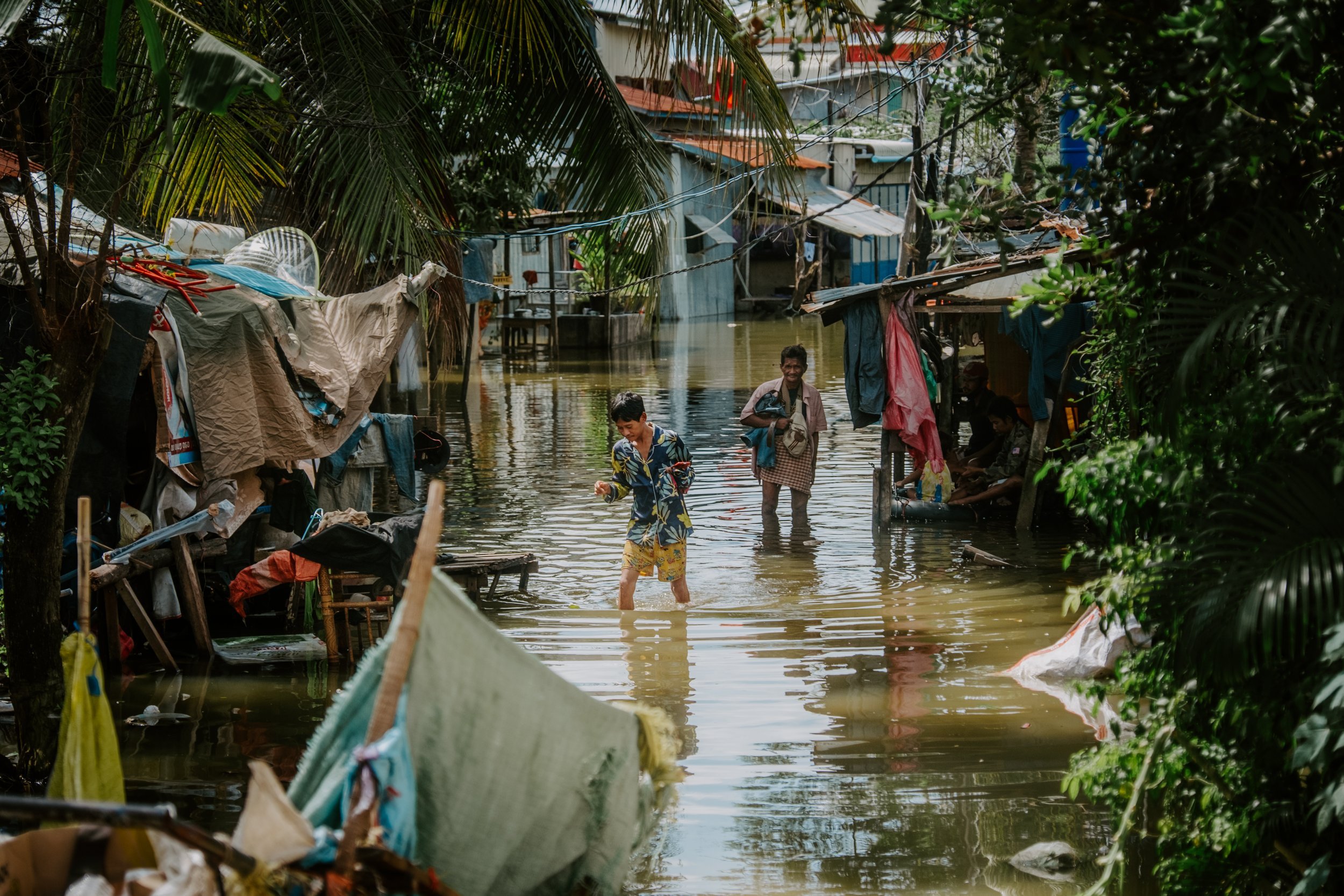 People wading through floodwaters in a residential area with makeshift houses, trees, and belongings submerged.