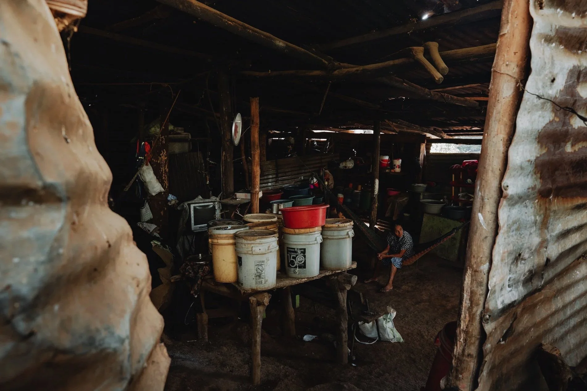 A woman sitting in a rustic, makeshift living space with wooden and metal walls, surrounded by buckets, containers, and household items.