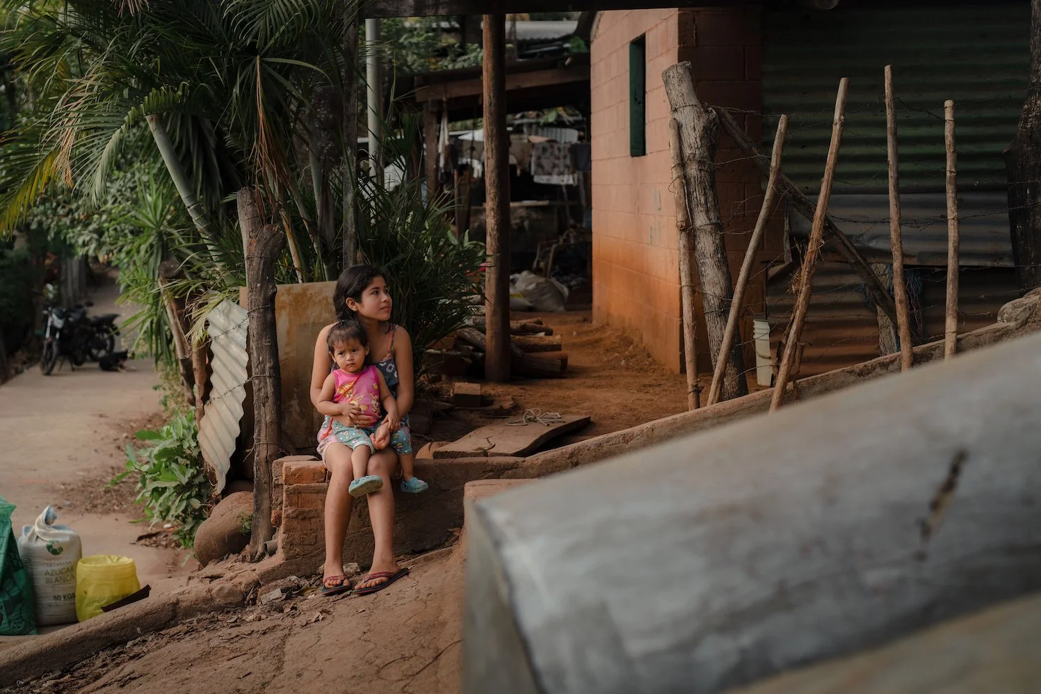 A young girl sitting on a small brick ledge holding a younger girl on her lap in a rustic outdoor setting with plants and makeshift fencing, near a corrugated metal and brick building.