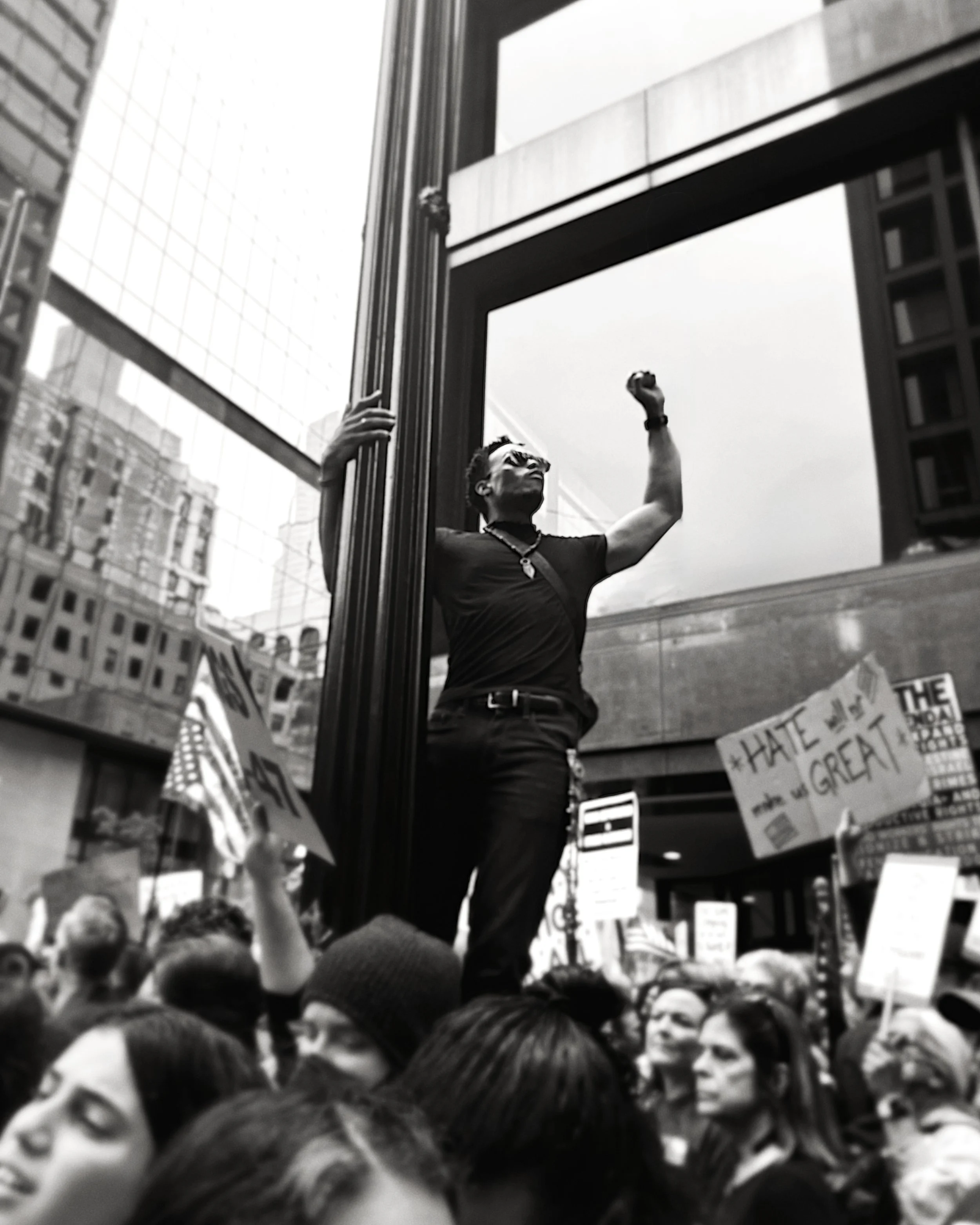 Black and white photo of a protest with a crowd, some holding signs, one partially visible sign says 'HATE' and 'GREAT,' and a person standing on a ledge or structure with one hand on a pillar and the other raised.