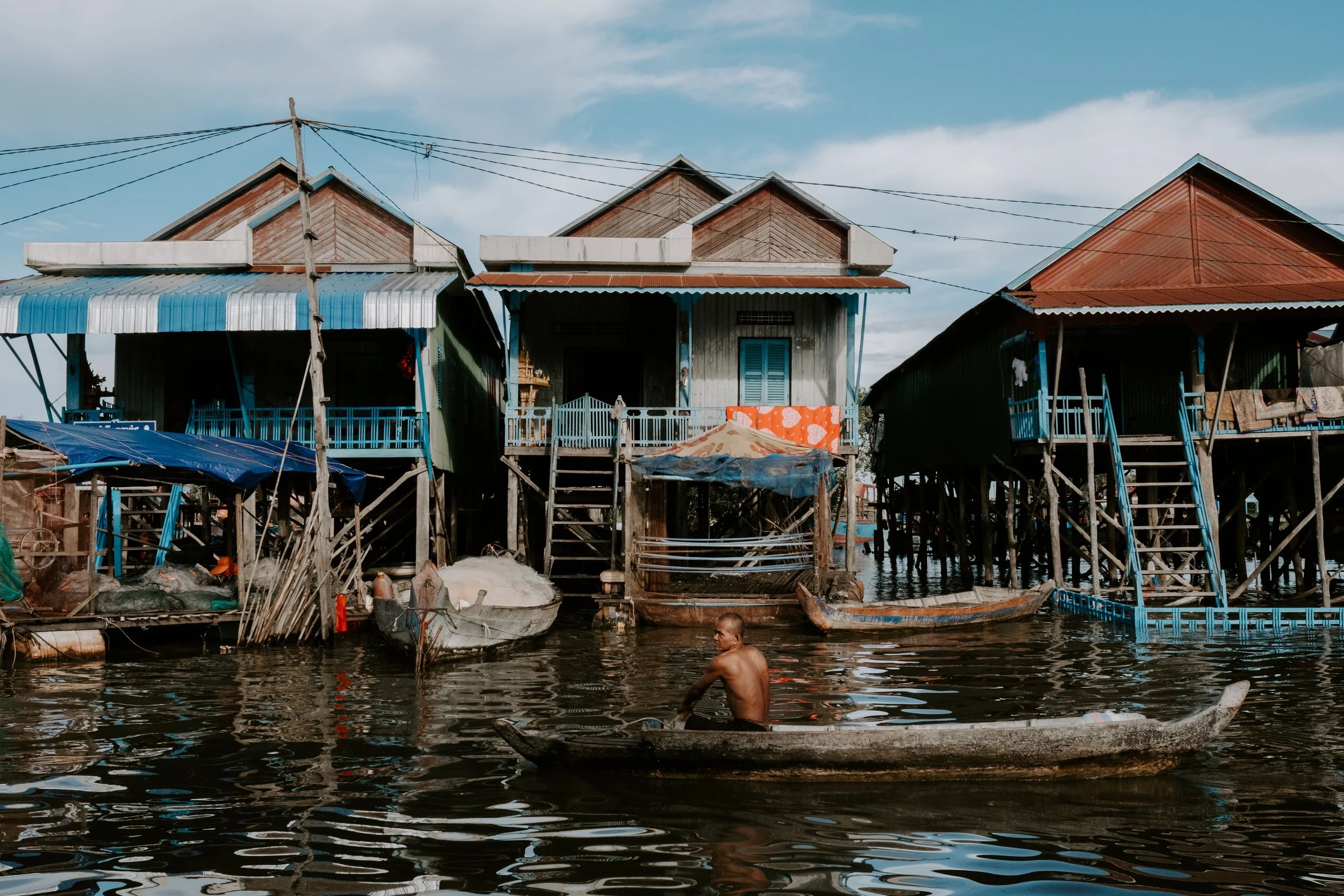 A man paddling a small boat on water in front of stilt houses with wooden and metal exteriors and stairs leading up to the entrances, under a partly cloudy sky.