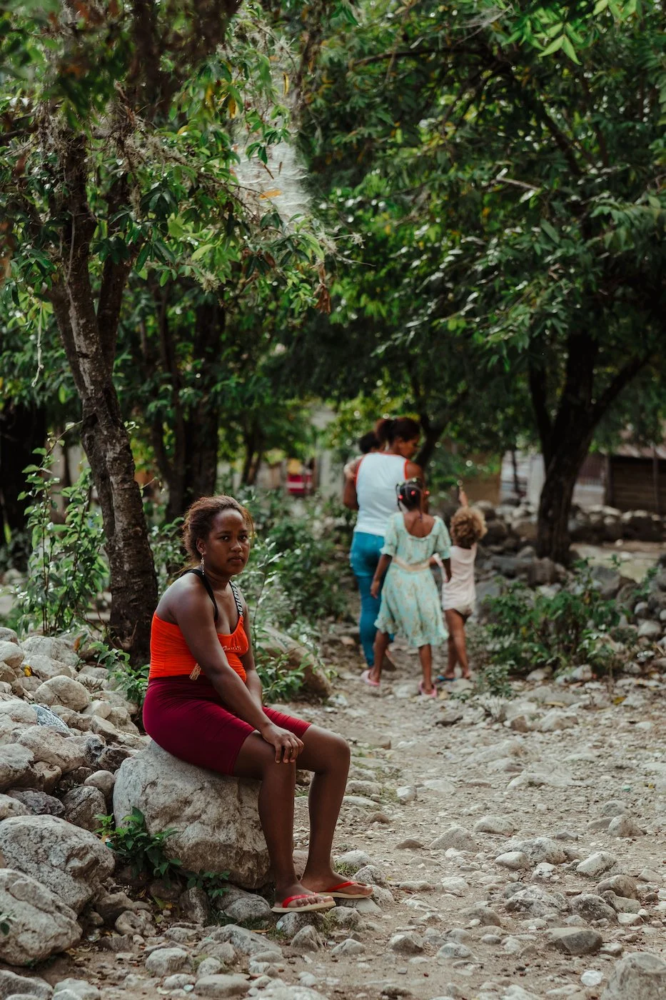 A young woman with curly hair sitting on a rock along a rocky dirt path surrounded by trees; in the background, a group of women and children walking along the path, with greenery and trees around.
