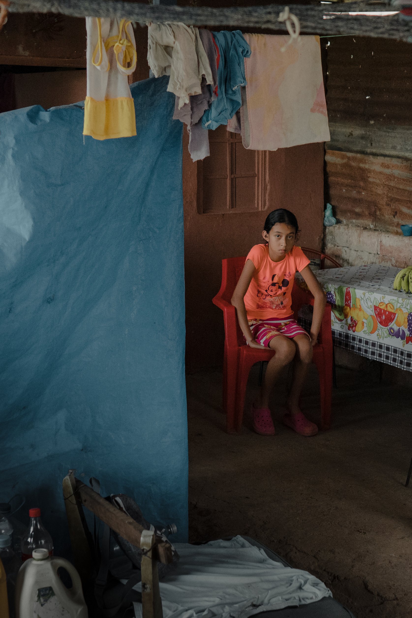 A young girl sitting on a red plastic chair inside a modest home with clothes hanging on a line overhead and laundry on the floor. A table covered with a colorful fruit-themed tablecloth is visible in the background.