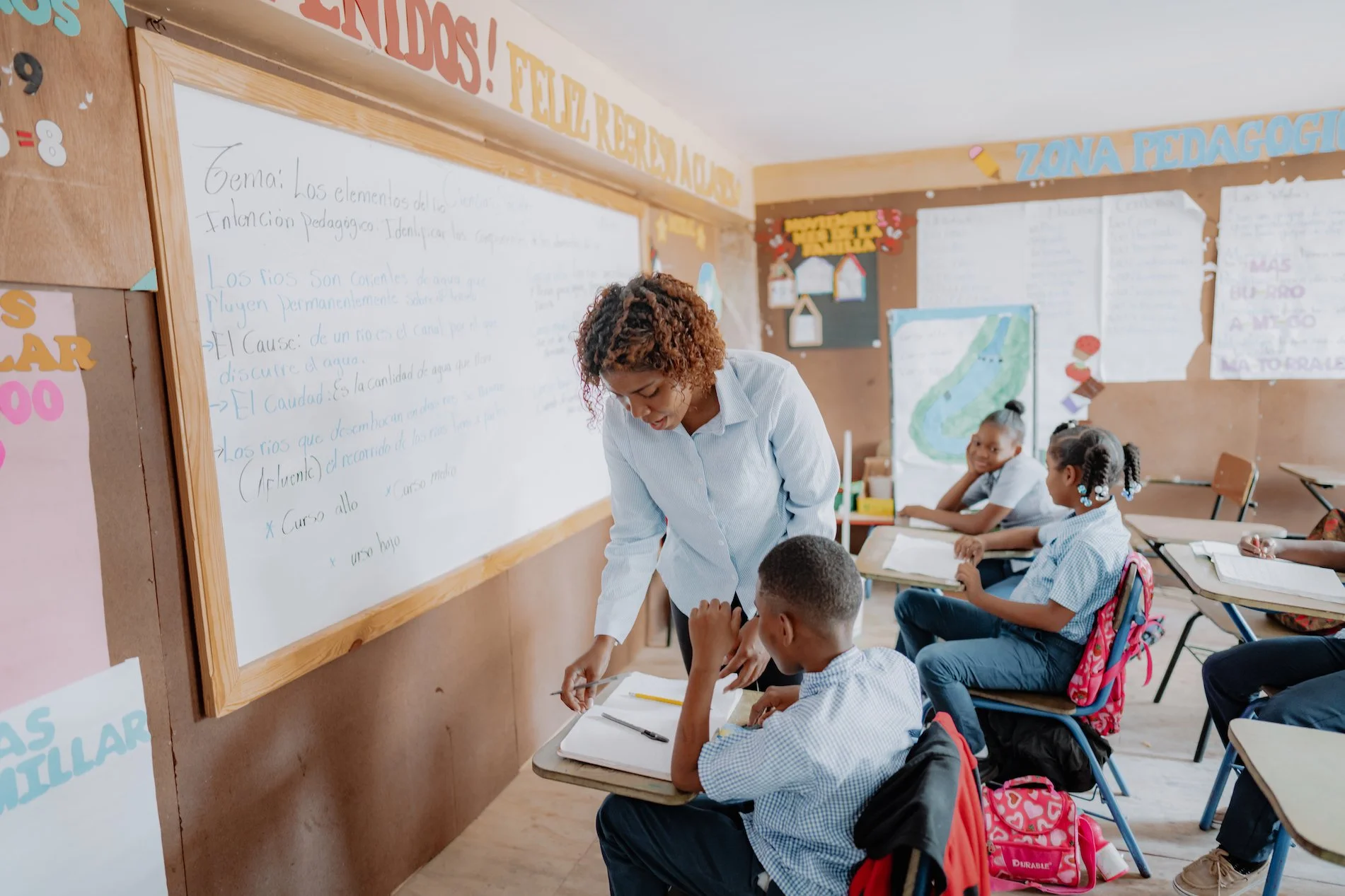 A classroom with a teacher assisting a student at his desk. Several students are seated at desks, paying attention. The classroom has educational posters and a whiteboard with writing.