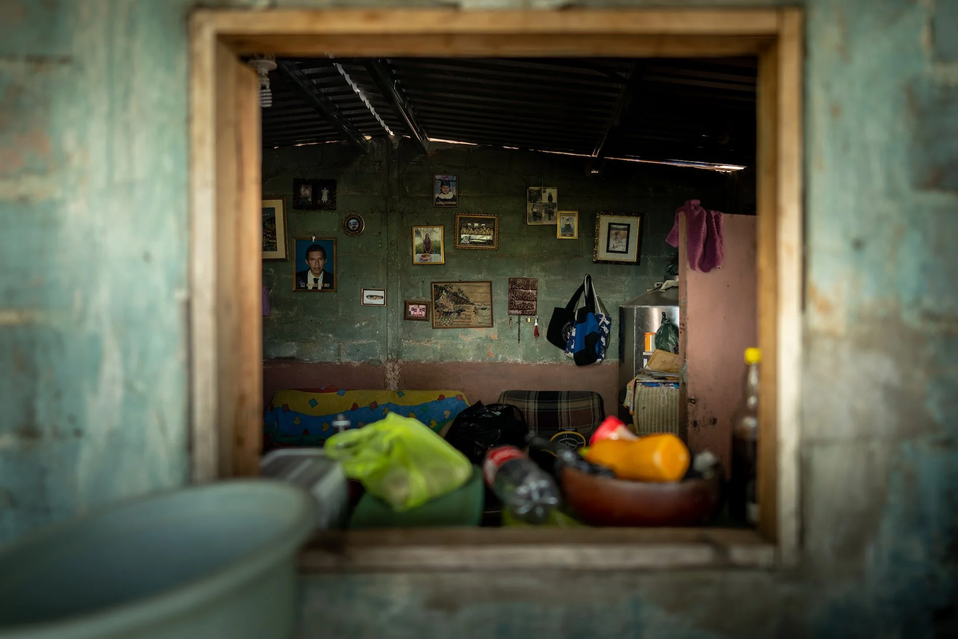 View of a room through a small window, showing a wall decorated with various framed photographs and artwork, a couch with colorful pillows, and various household items and bags inside.