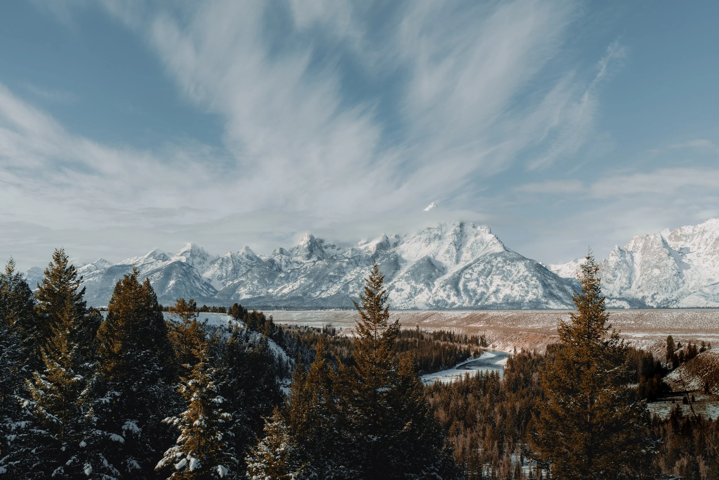 Snow-covered mountains with a partly cloudy sky, a river, and pine trees in the foreground.