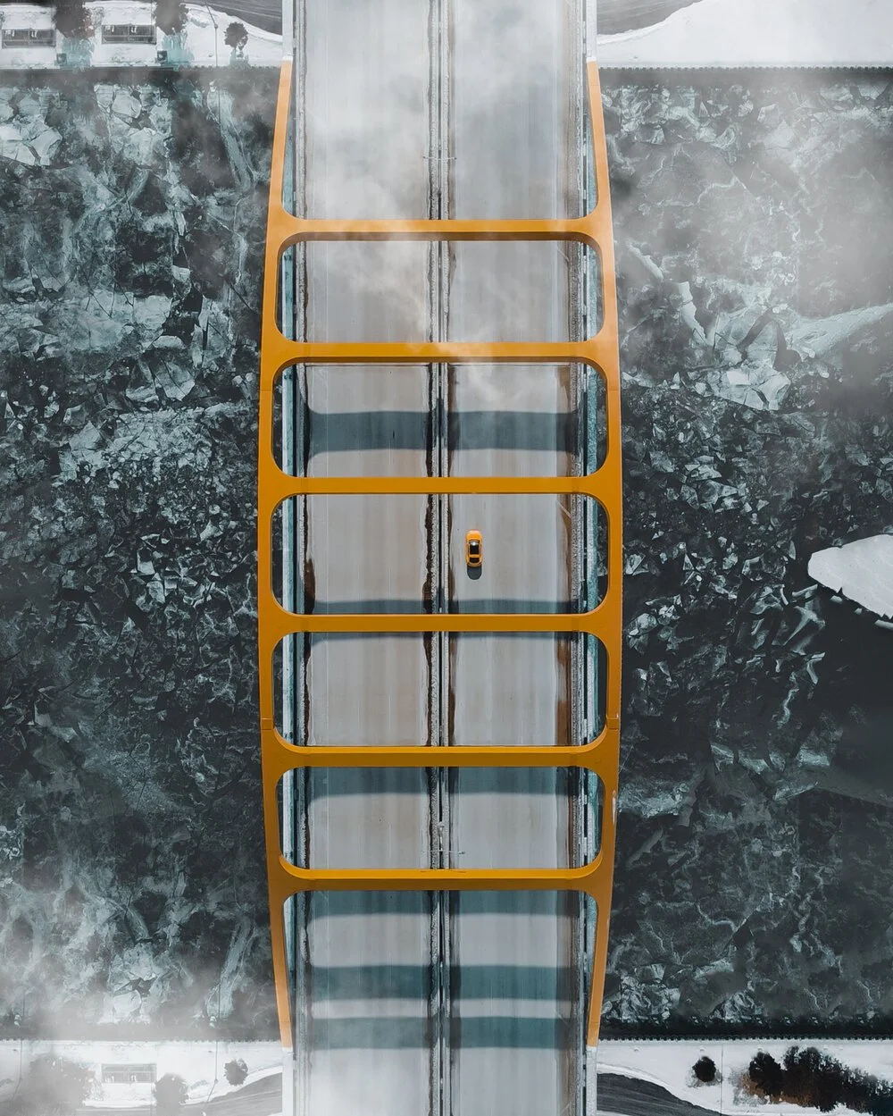 Aerial view of a yellow bridge with a single car crossing over a snowy, rocky river or canyon.
