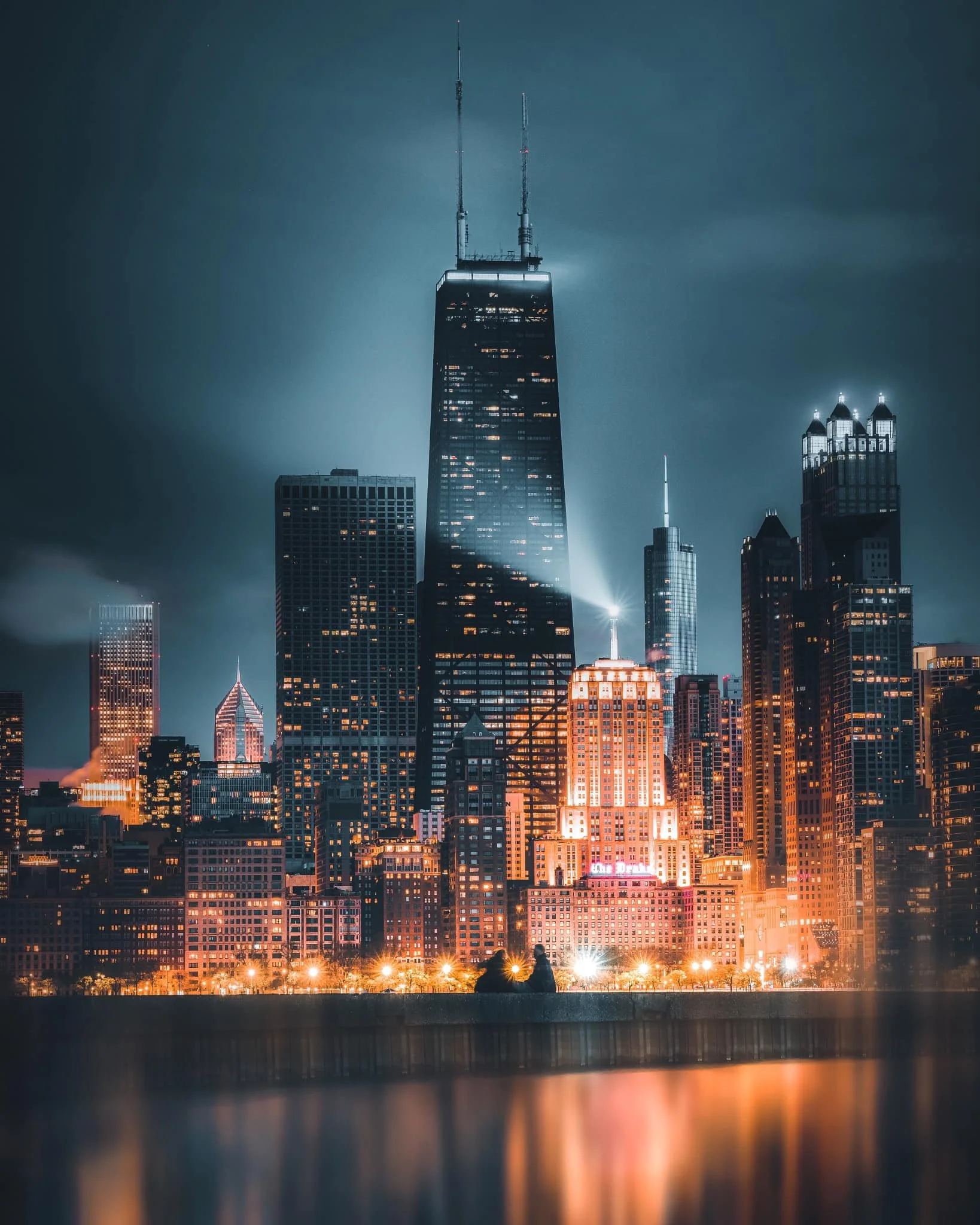 Nighttime view of a city skyline with tall illuminated buildings reflected on a body of water, with two people sitting on a bench in the foreground.