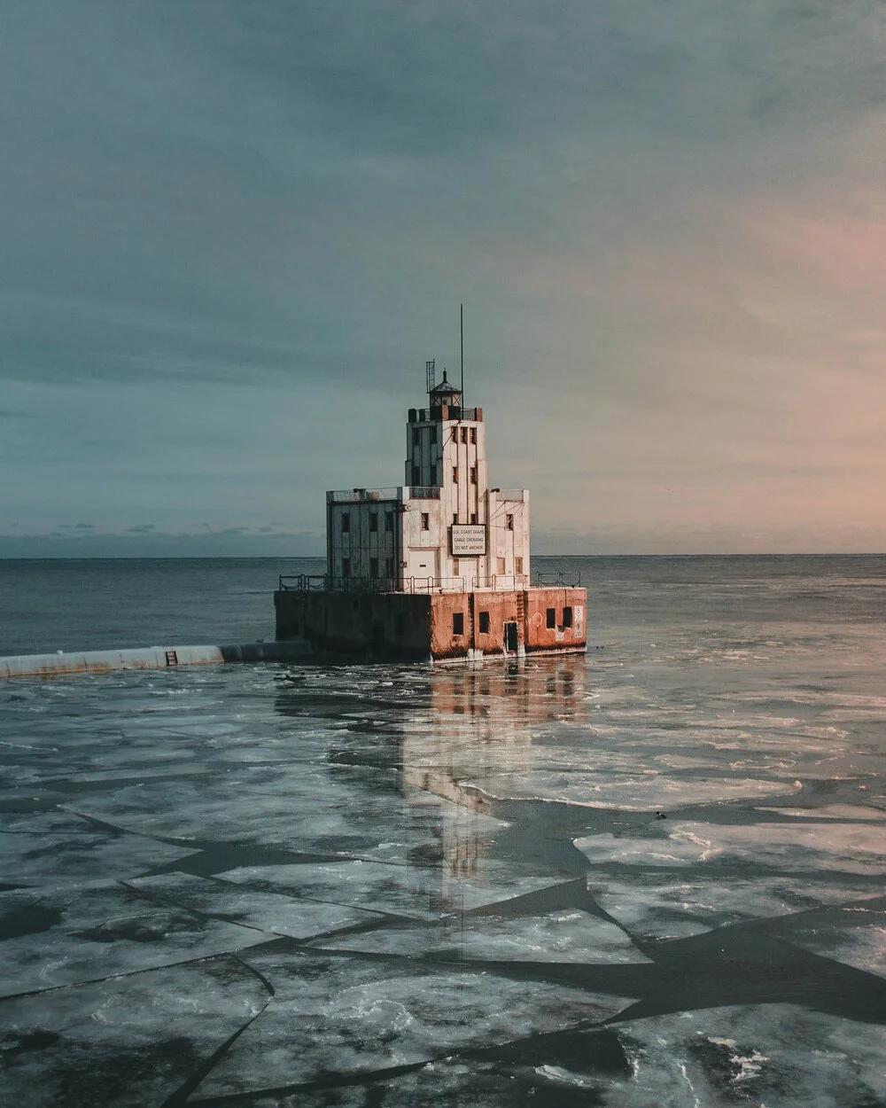 A lighthouse on a rocky, icy shoreline with a calm sea and a cloudy sky at sunset or sunrise.