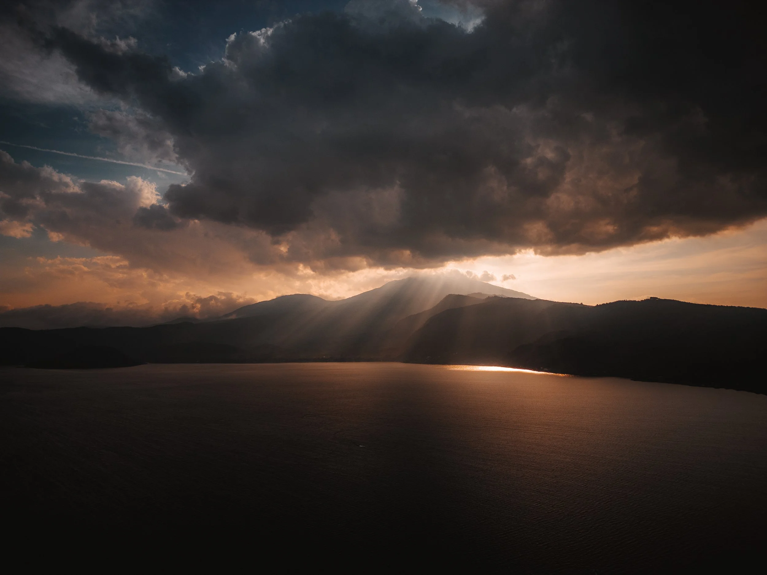 Sun rays breaking through dark clouds over a mountain range and a body of water at sunset.