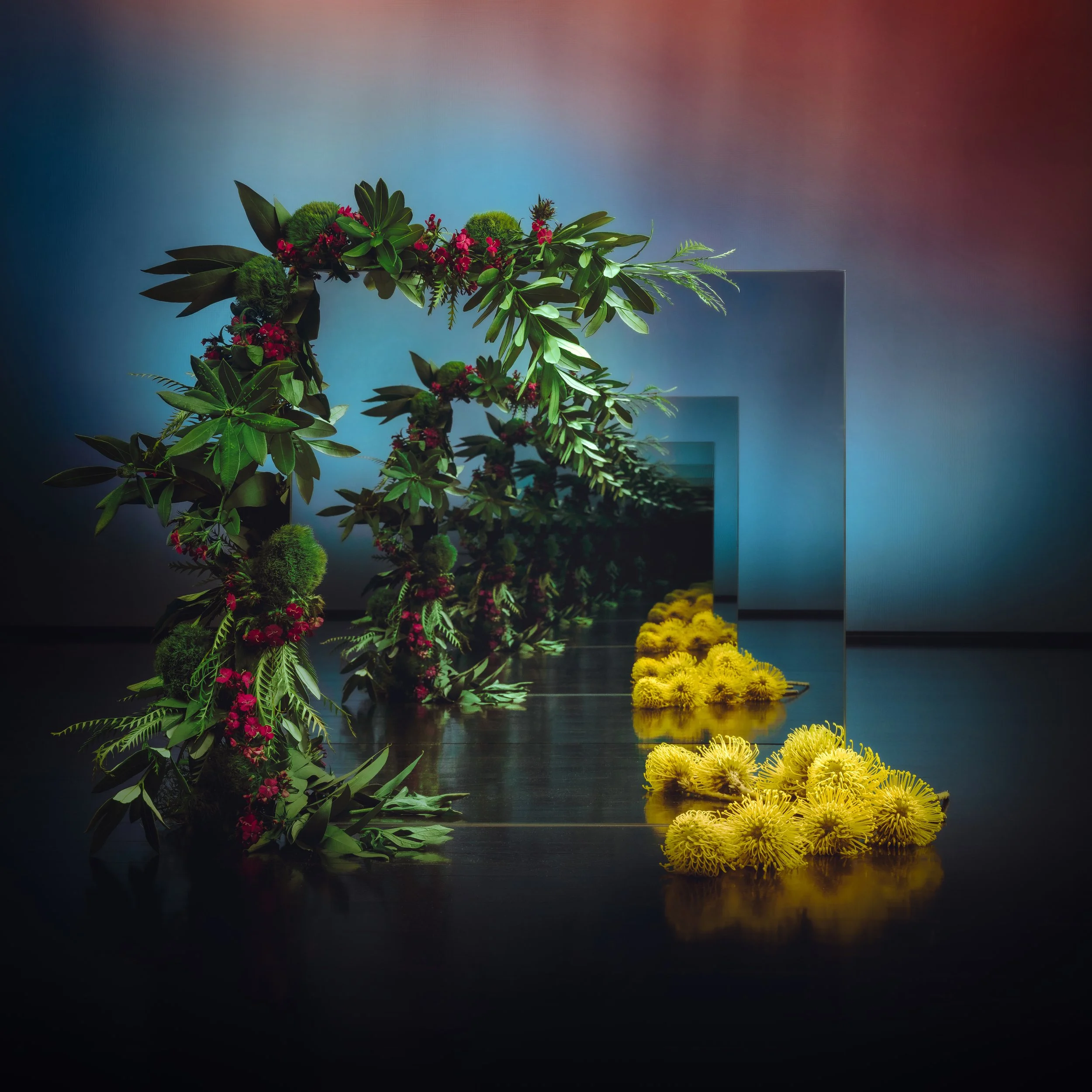 Arrangement of flowers and greenery forming a circular arch with a mirror reflection, accompanied by yellow flowers on the floor, set against a gradient background.