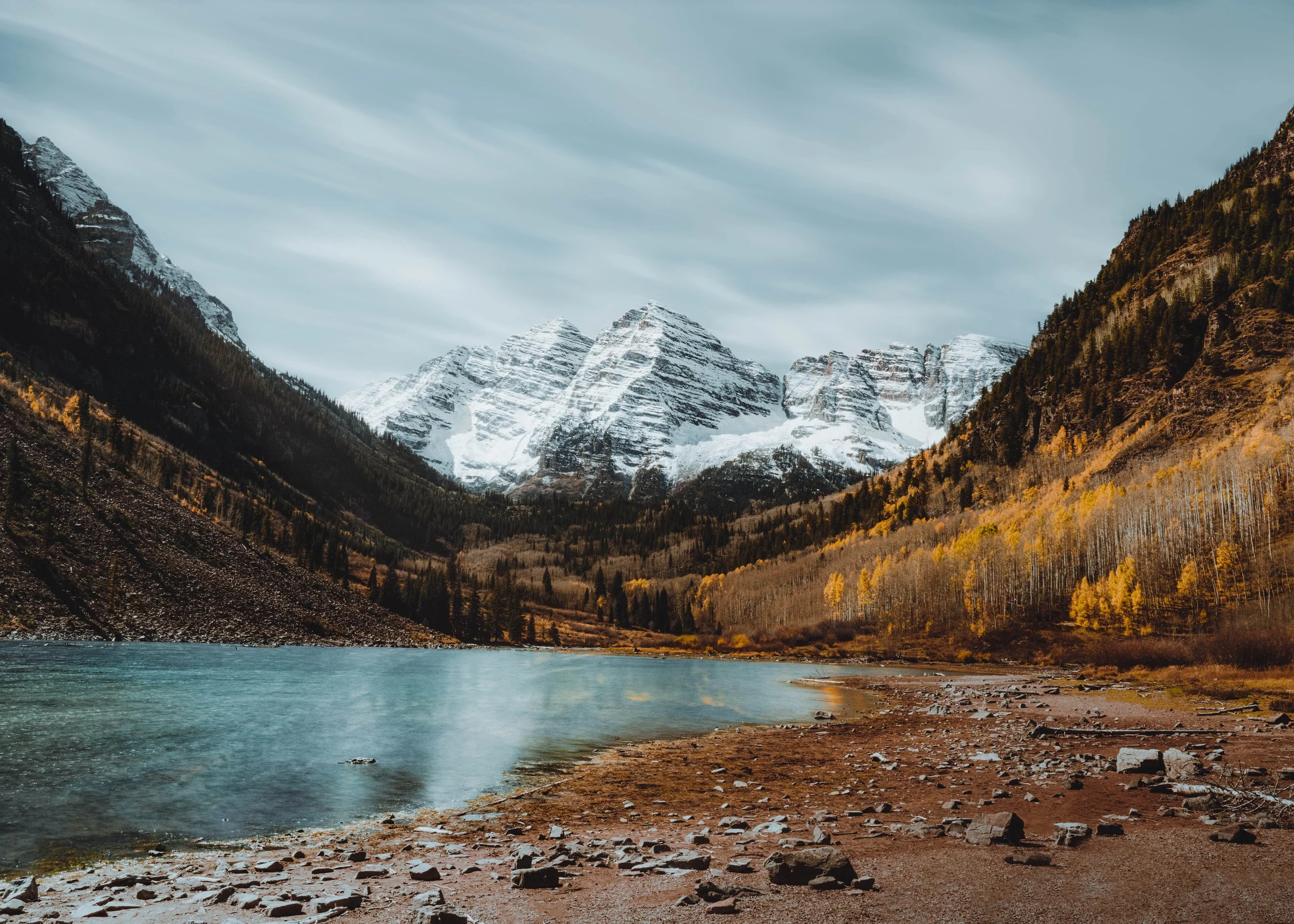 A scenic view of a mountain landscape with snow-capped peaks, a river in the foreground, and autumn trees on the slopes.