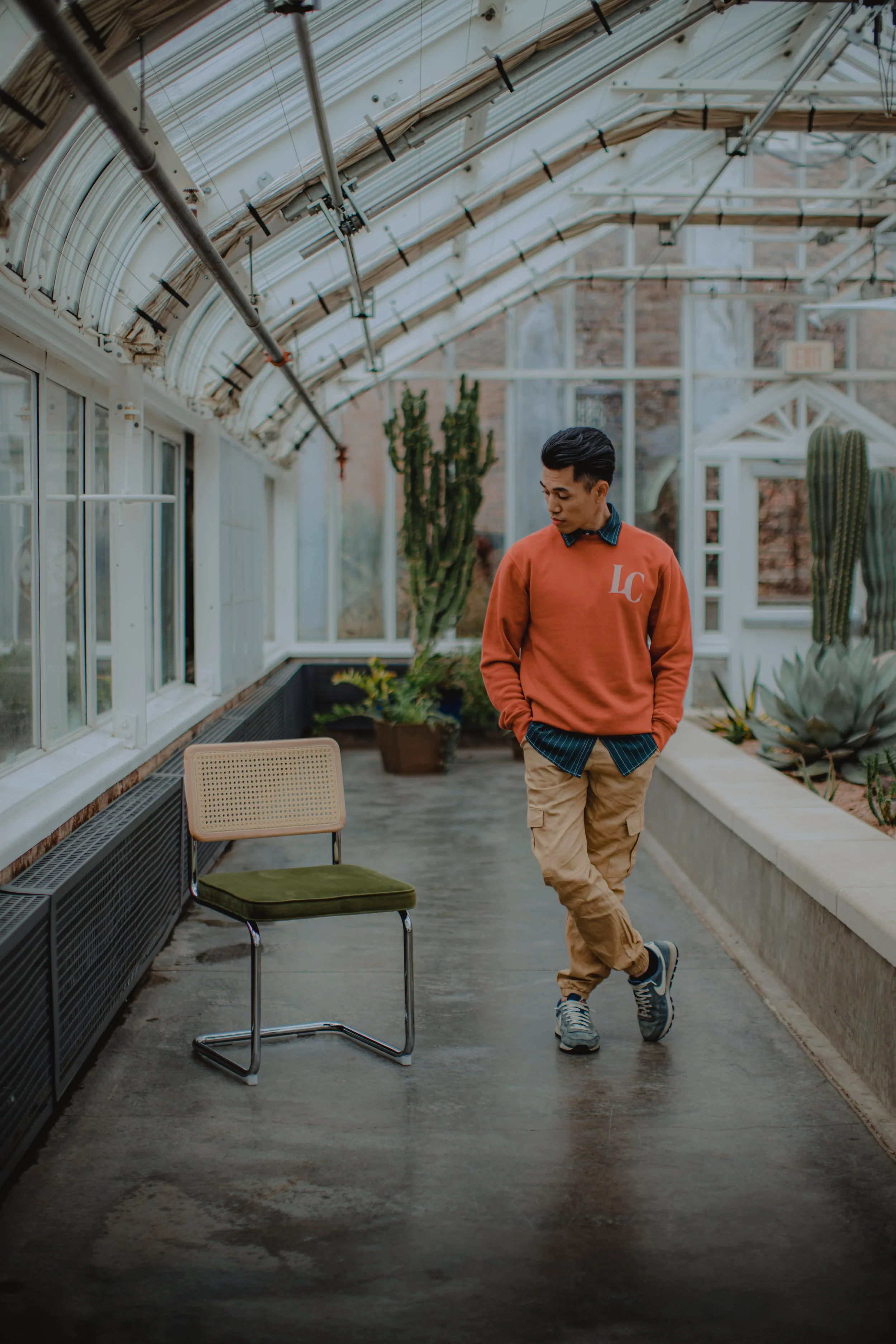 A young man in a red sweatshirt, beige cargo pants, and sneakers stands inside a greenhouse with glass walls and ceiling, surrounded by various cacti and succulent plants, with an empty chair nearby.