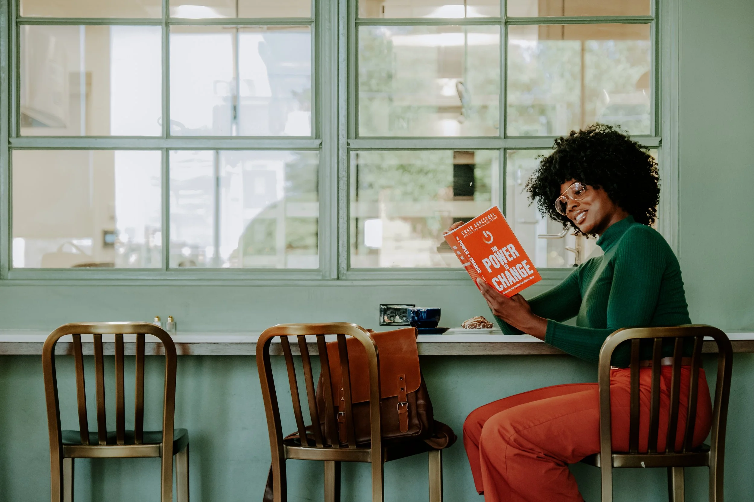 A woman with curly hair and glasses sitting at a counter reading a book titled "The Power of Change" in a bright cafe or restaurant with green walls and large windows.