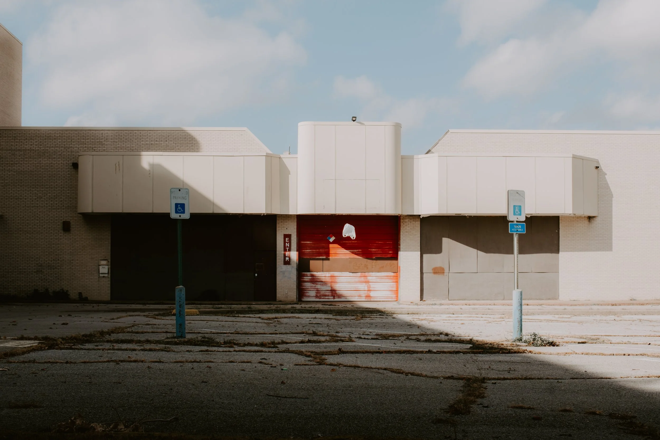 An empty parking lot in front of an abandoned building with a red rolling door and two disabled parking signs.