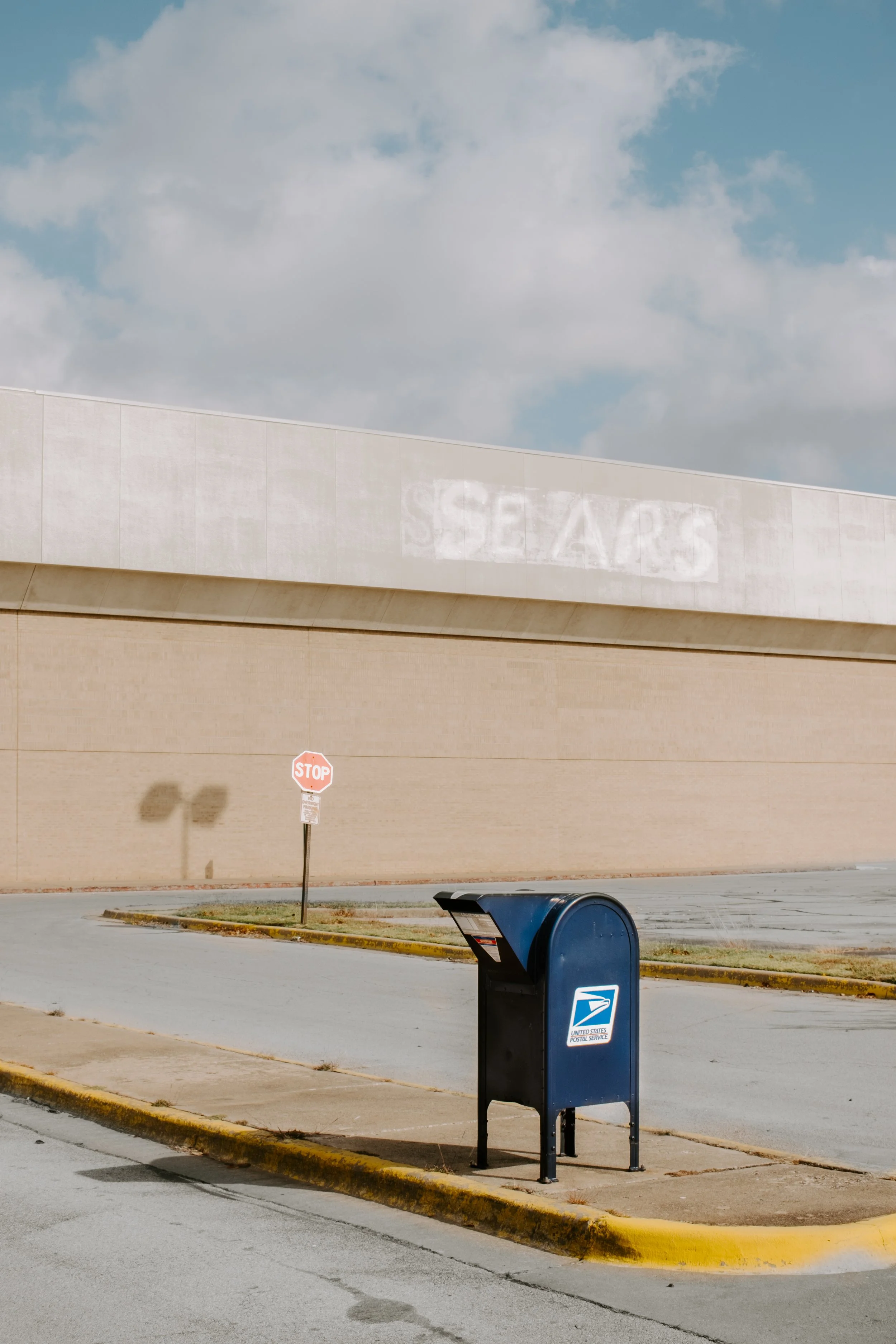 A USPS mailbox on a sidewalk in front of a beige brick wall with a stop sign and a large white building with a faded sign that reads 'SEARS' against a cloudy blue sky.