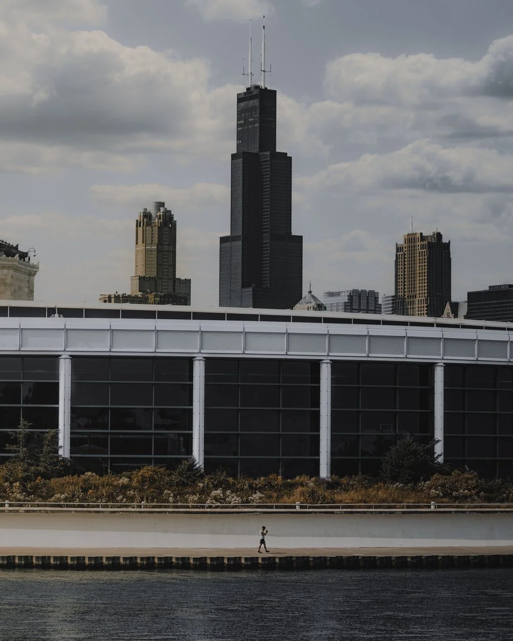 A person jogging along a waterfront with Chicago skyline in the background, featuring the Willis Tower and other tall buildings under a partly cloudy sky.
